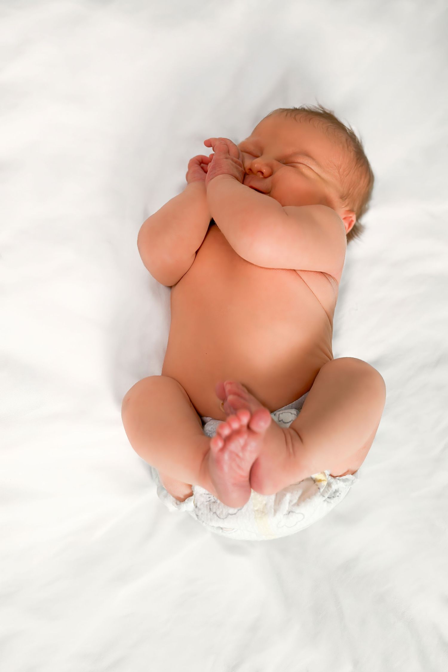 Newborn baby sleeping peacefully on white blanket wearing diaper with hands near face and curled legs.