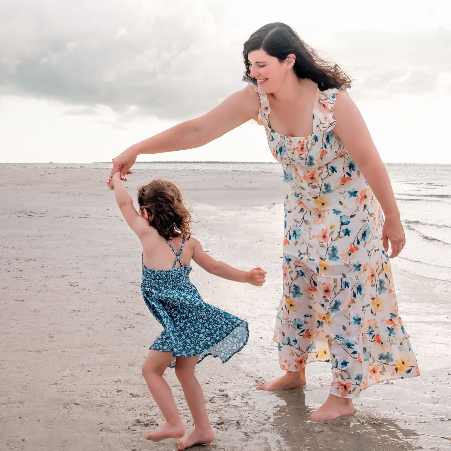 A mother and young daughter dance together on a sandy beach, both wearing floral dresses, with ocean waves in the background.