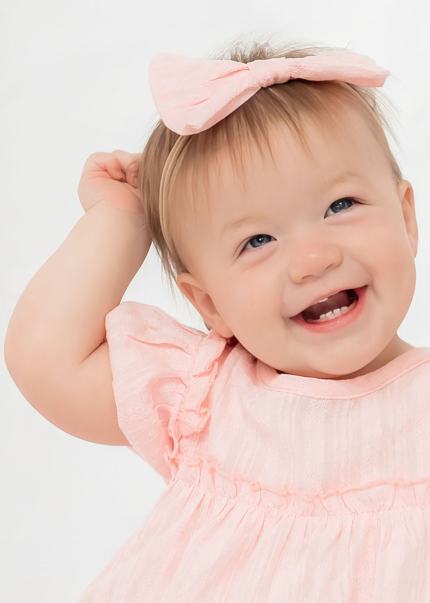Smiling baby girl in pink dress and bow headband, showing first teeth, on white background.
