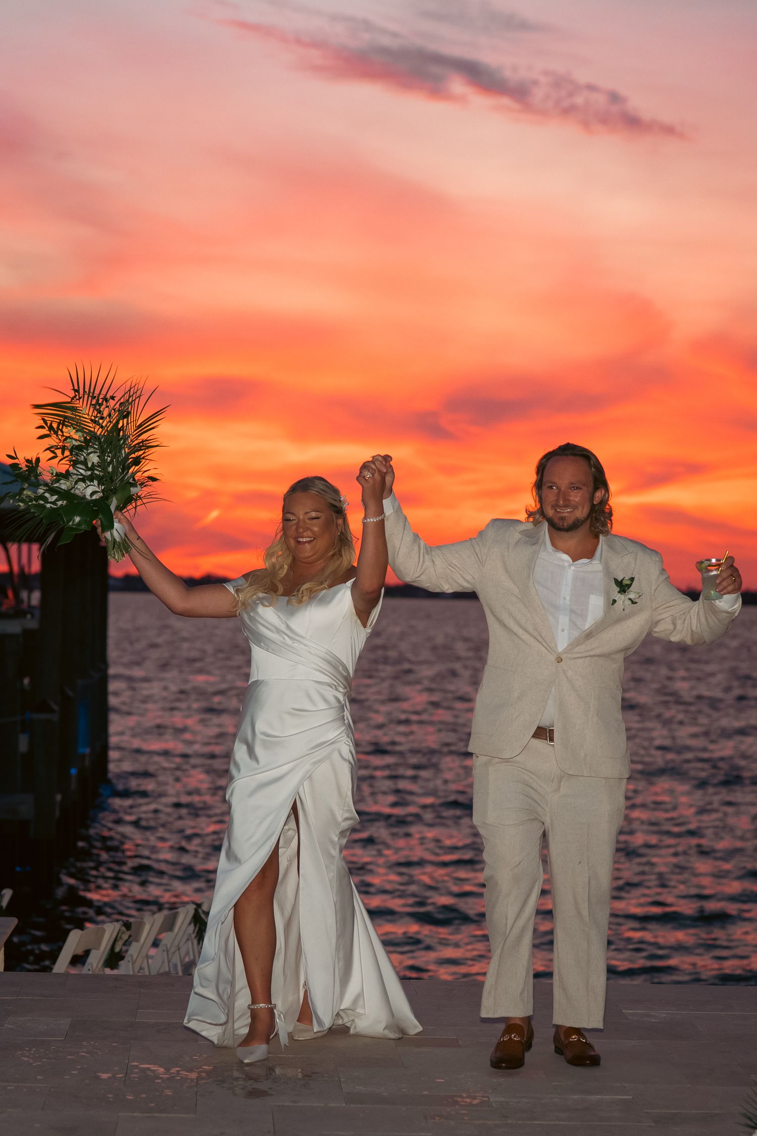 Bride and groom celebrate at sunset wedding ceremony by the ocean, raising hands joyfully under a vibrant red sky.