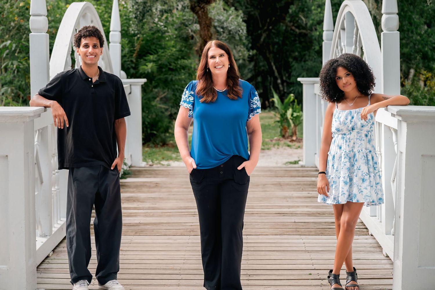 Three people posing on a white wooden bridge surrounded by lush green trees on a sunny day.