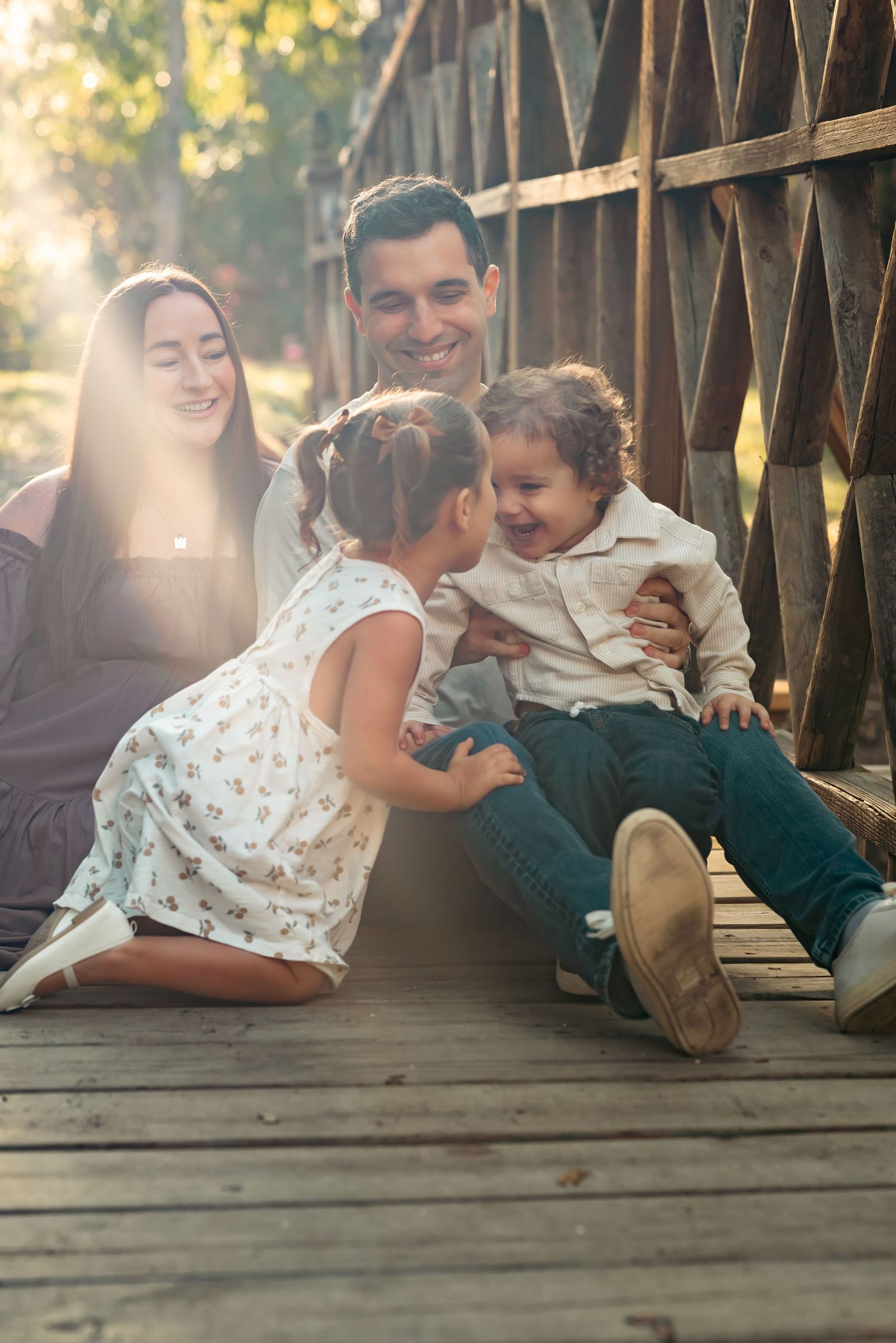 Happy family sitting on wooden bridge with two young children laughing and playing in warm sunlight.