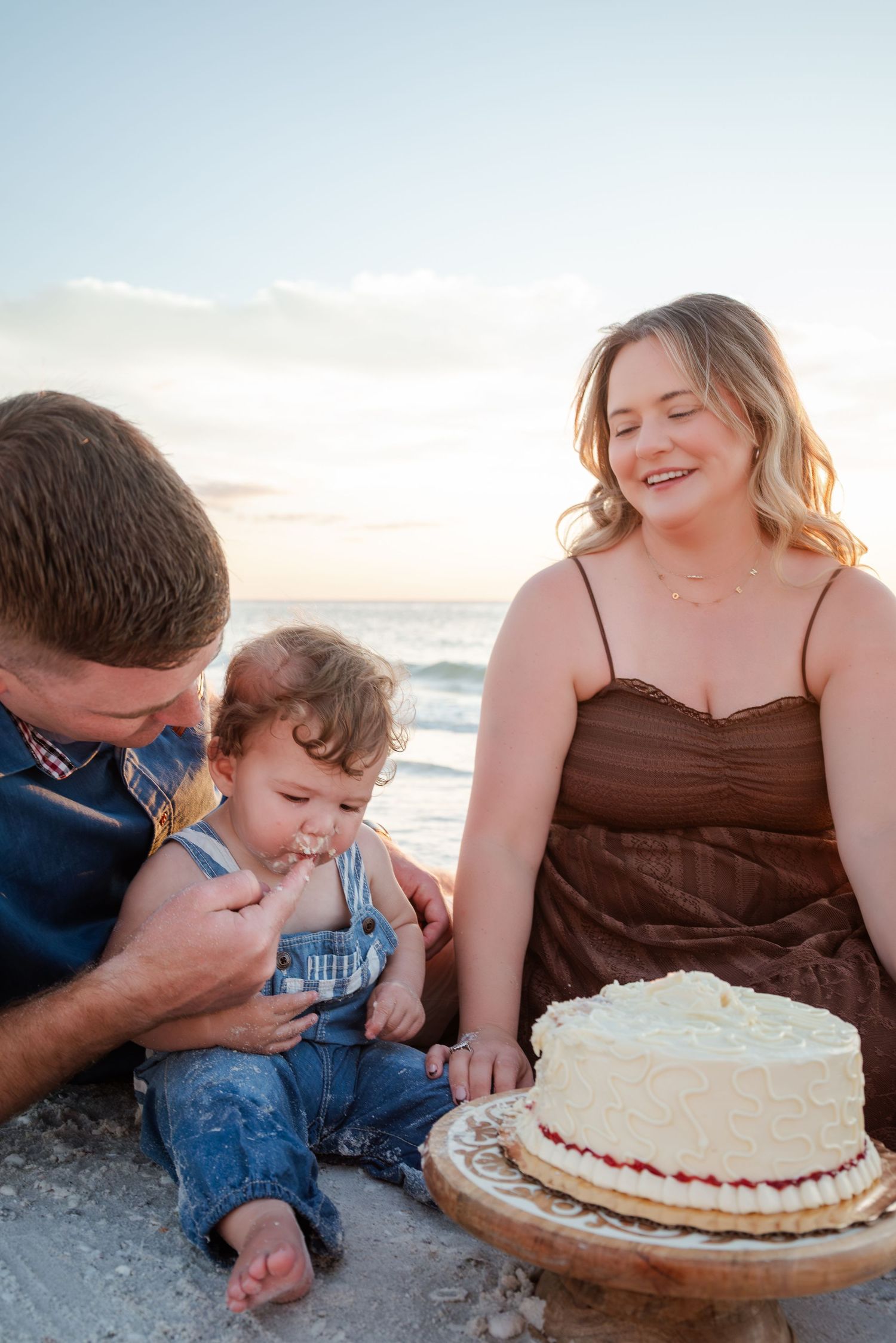 Young parents smiling as their baby enjoys first birthday cake on a sandy beach at golden hour.