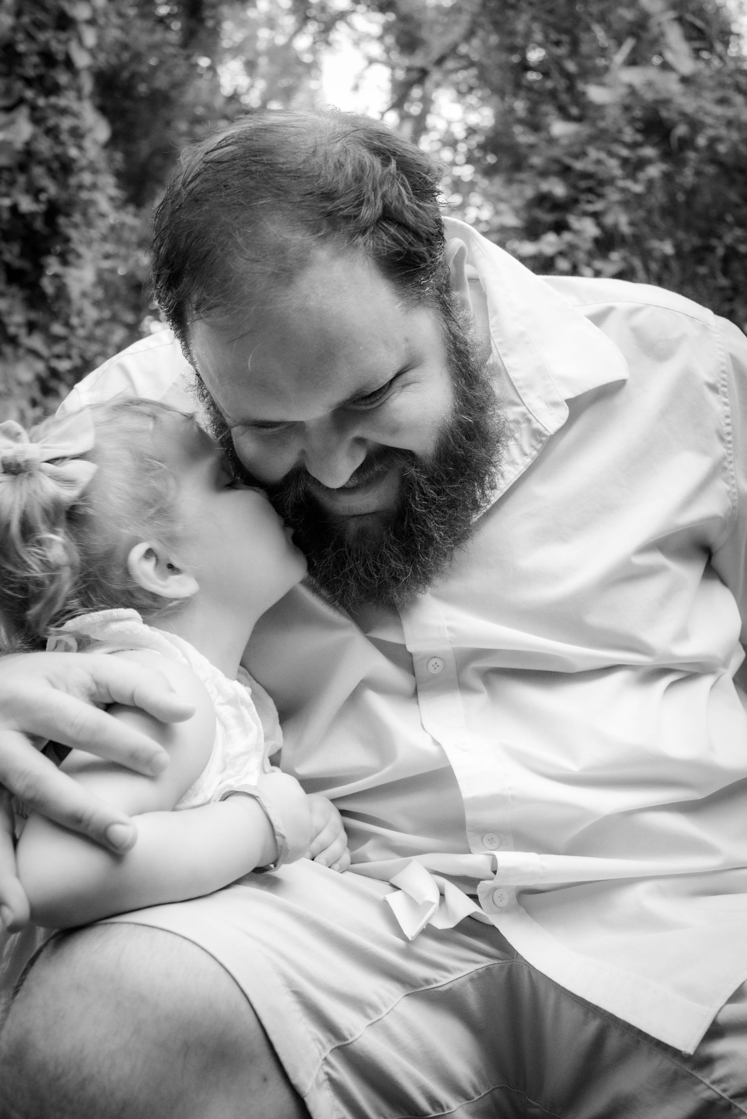 Black and white photo of bearded father laughing as baby kisses his cheek outdoors.