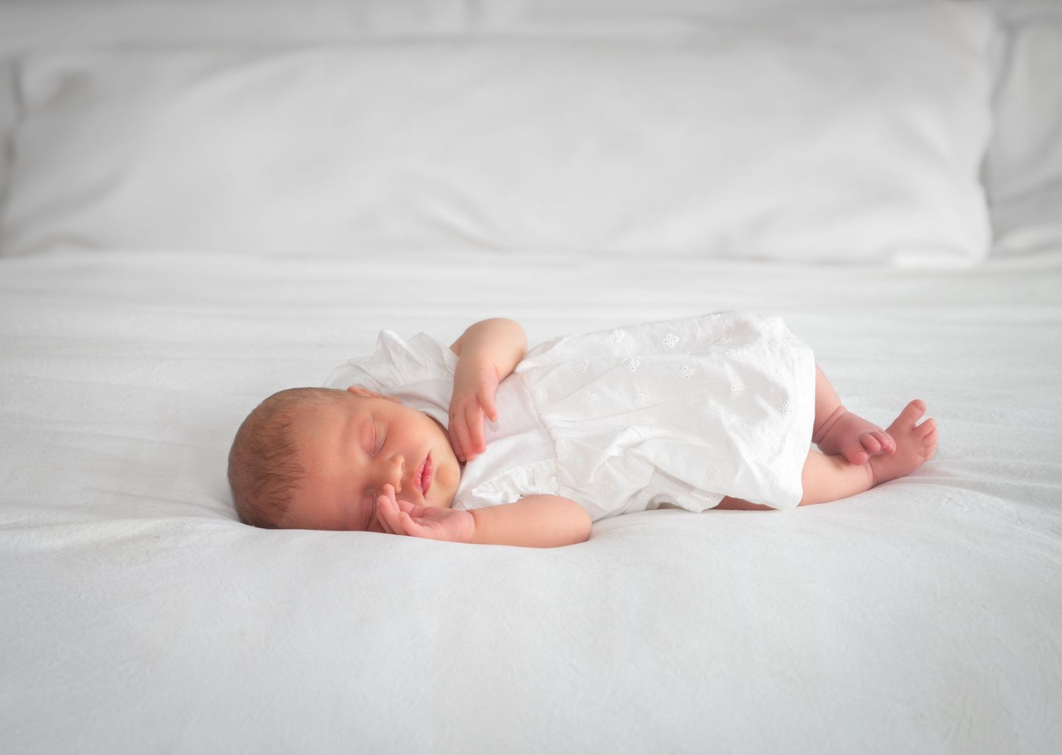 Peaceful newborn baby sleeping on white bedding wearing a white onesie.