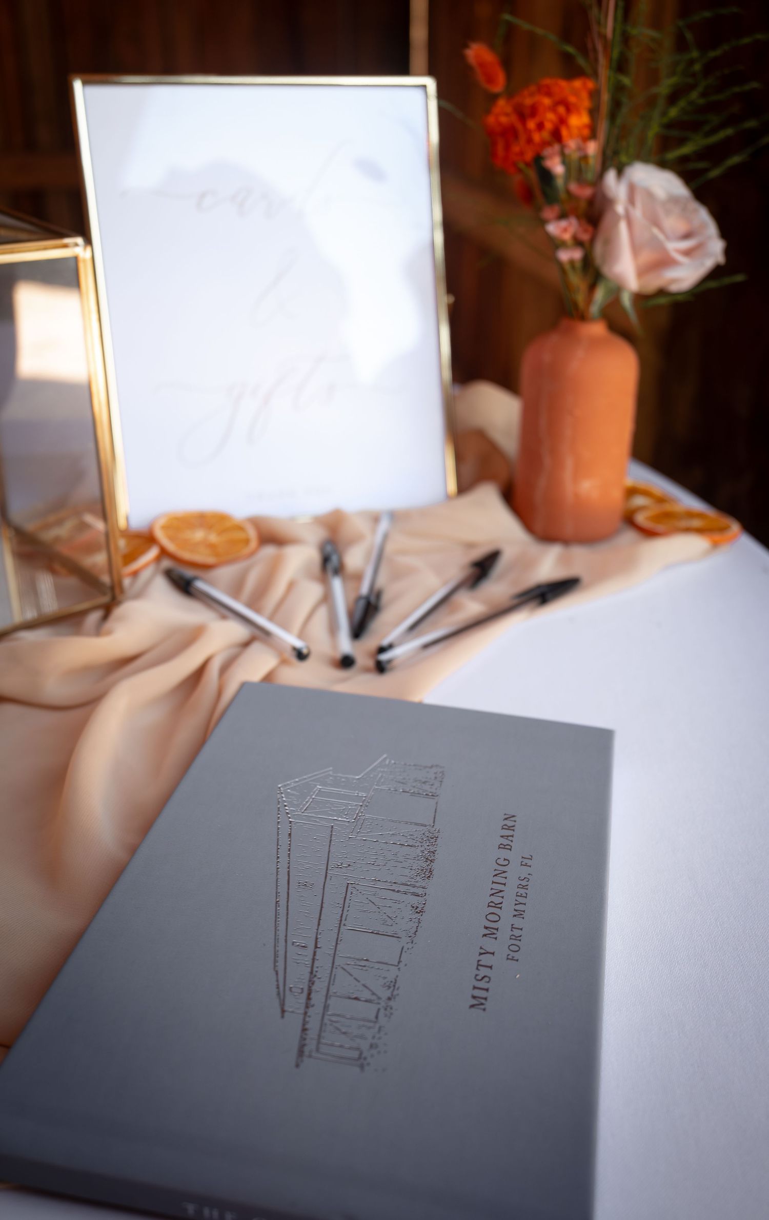 Elegant wedding guest book display with terracotta vase, orange flowers, dried citrus slices, and framed monogram sign.
