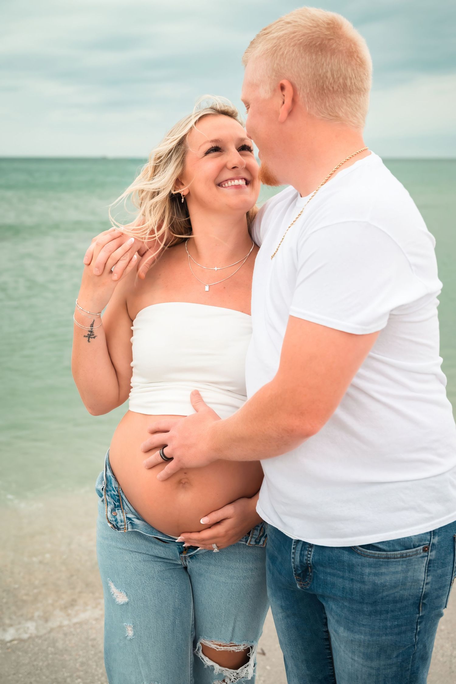 Smiling pregnant woman in white tube top and jeans embraced by partner at ocean shore.