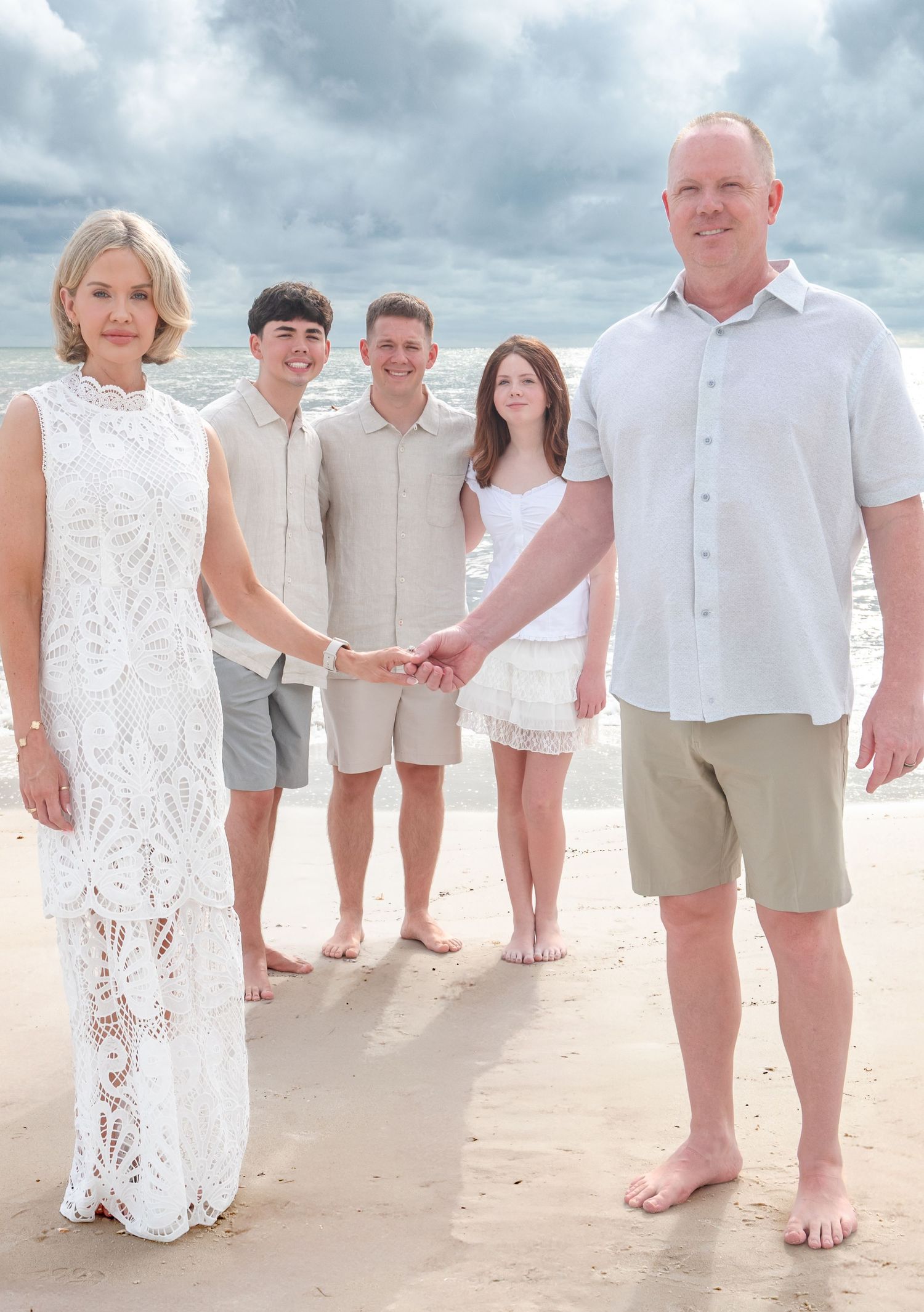 Happy family of five posing together on a sandy beach with cloudy skies, dressed in coordinating white and beige outfits.