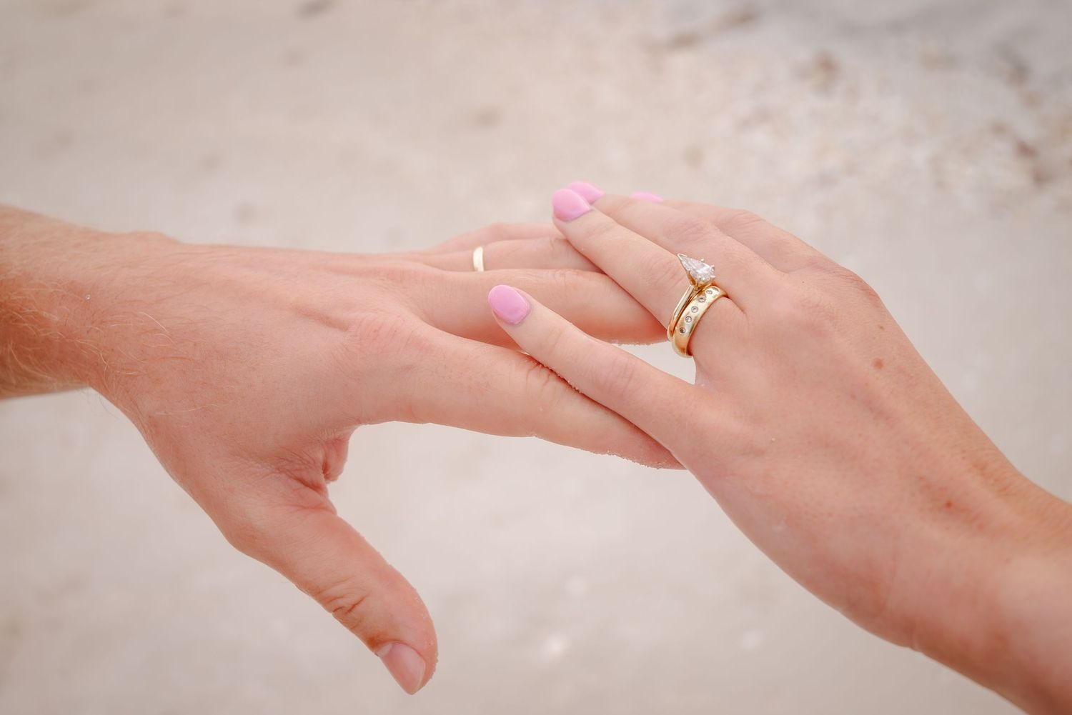 Close-up of two hands touching, both wearing gold wedding bands, with pink manicured nails visible.