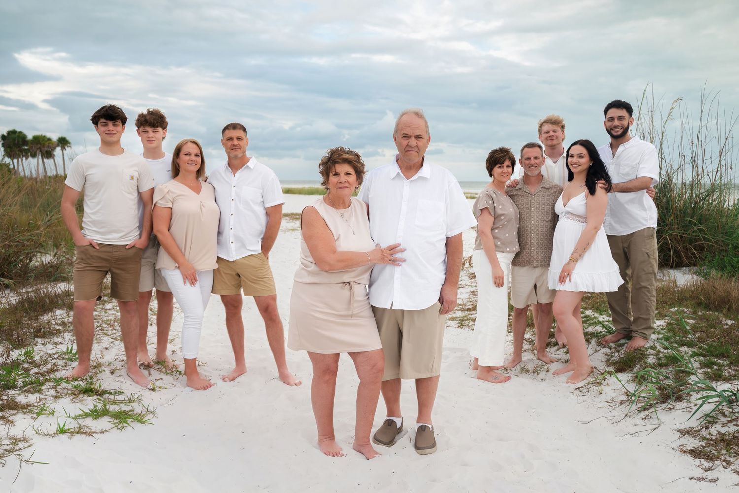 Family portrait of multigenerational group in coordinated beige and white outfits on a sandy beach.