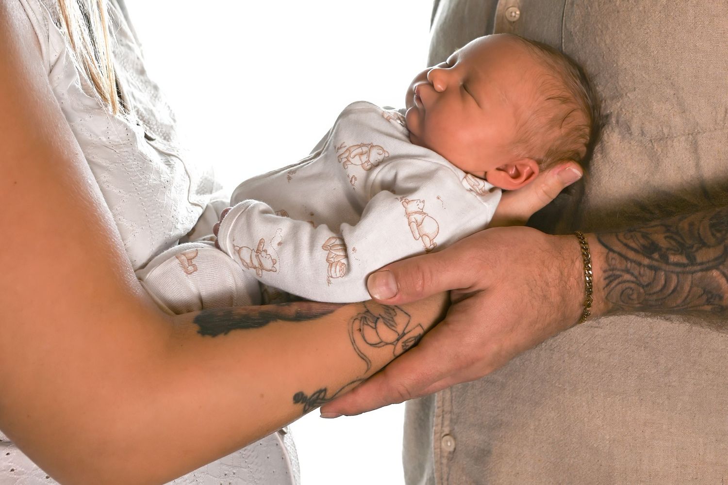 Tattooed parents cradling a sleeping newborn baby dressed in a white onesie with delicate prints.