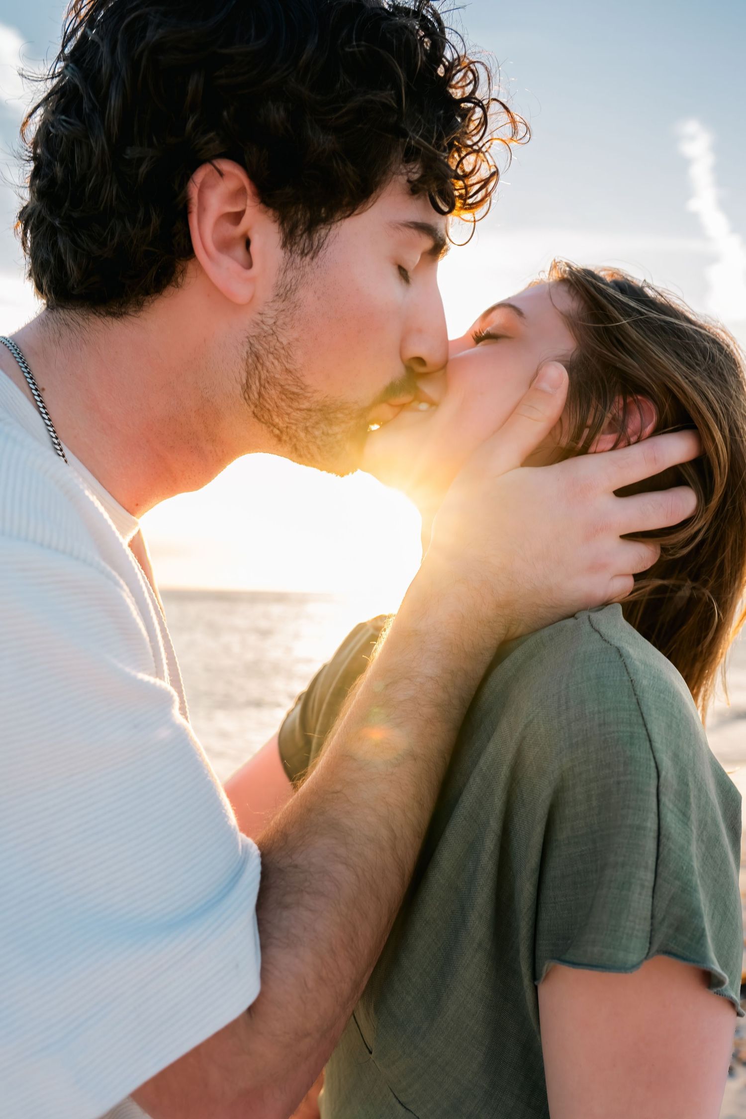 Couple sharing a romantic sunset kiss by the ocean, man gently holding woman's face in warm golden light.