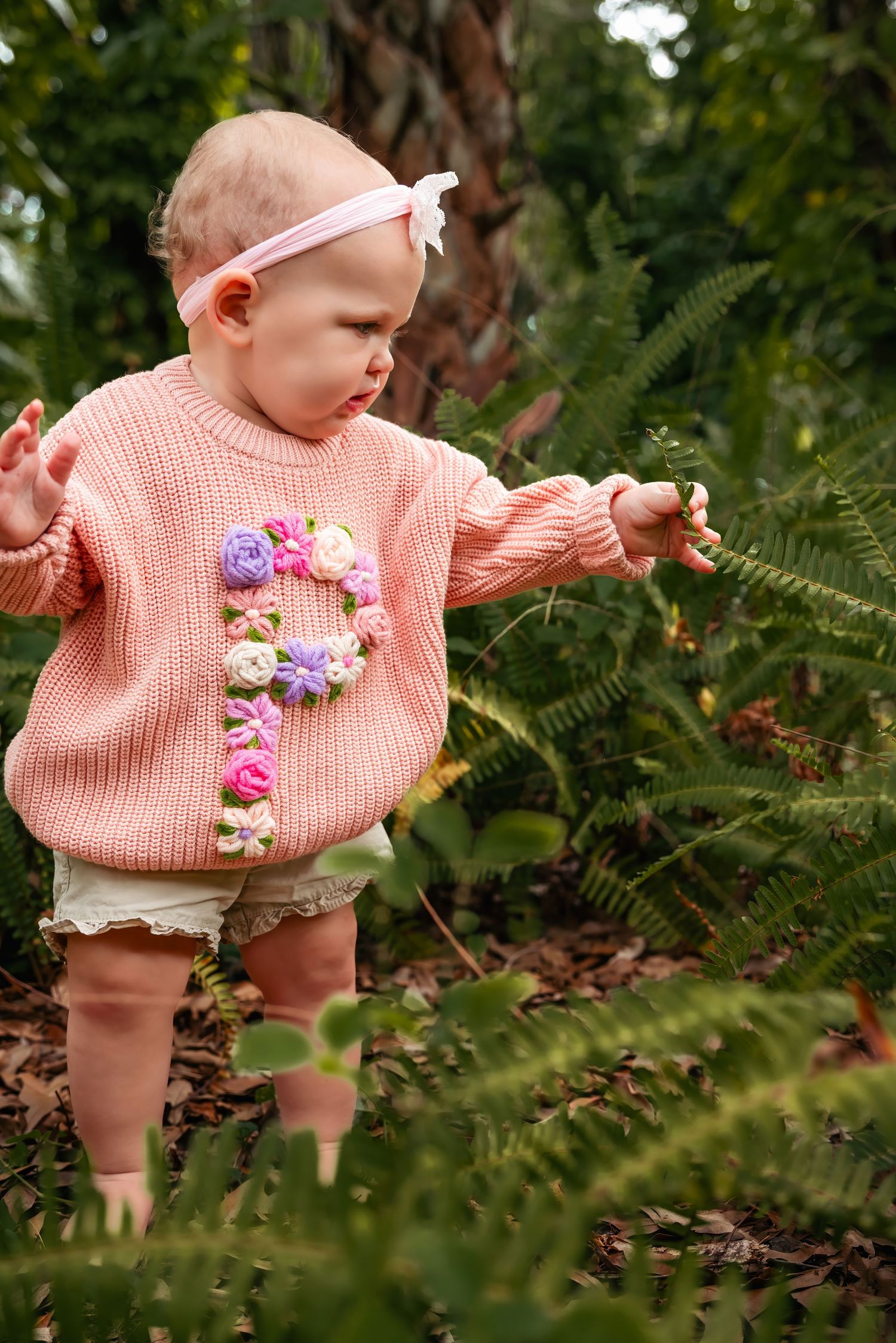 Baby girl in pink floral knit sweater exploring ferns in a garden, wearing a pink headband.