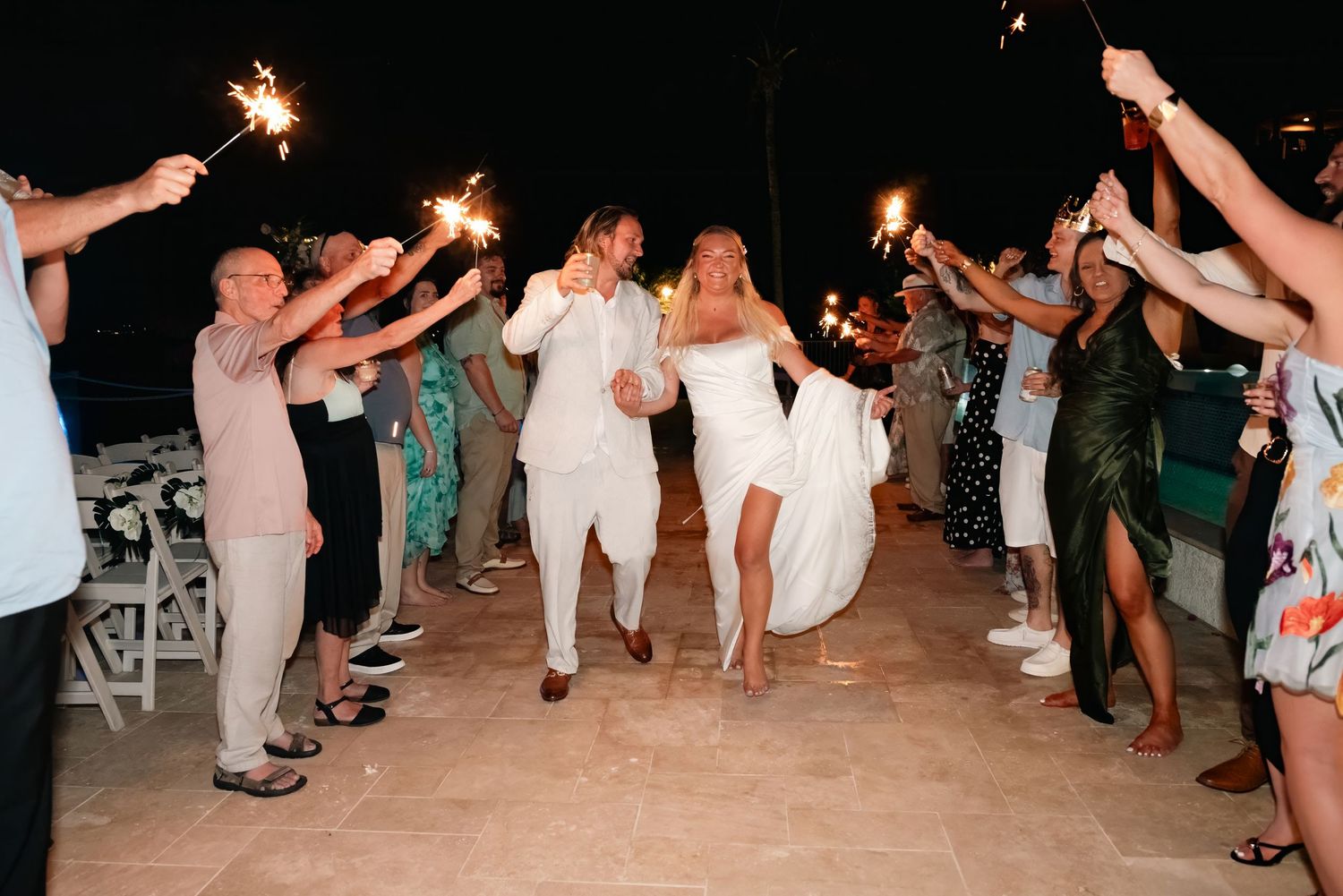 Bride and groom exit wedding reception through sparkler tunnel surrounded by cheering guests at night.