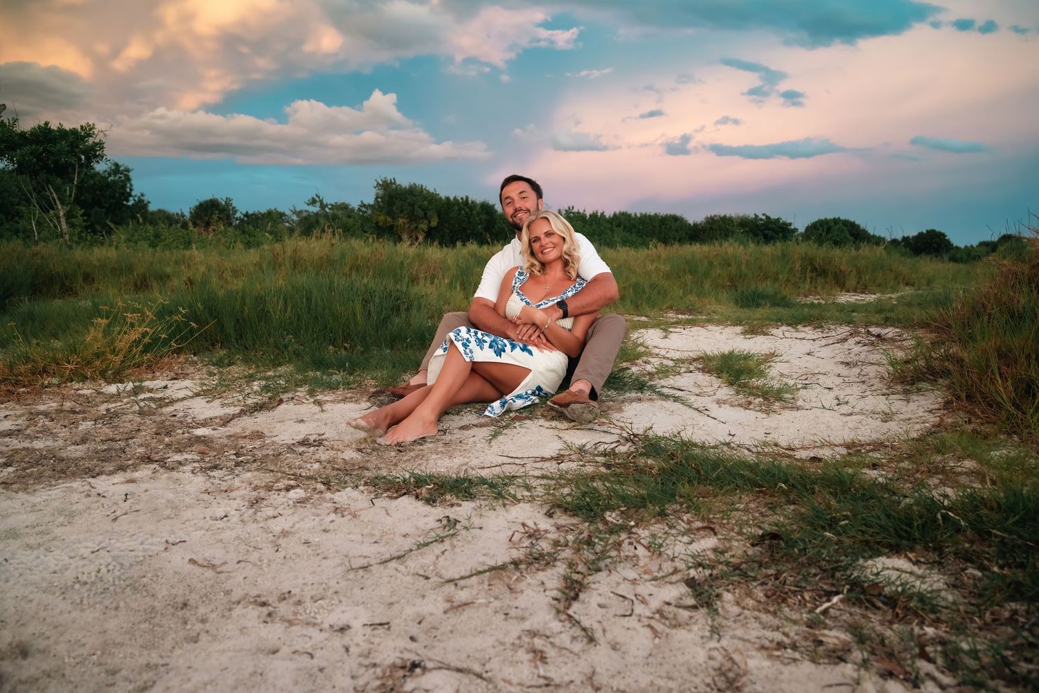 Couple sitting together on sandy beach at sunset, woman in blue dress leaning against man in white shirt, lush greenery behind.
