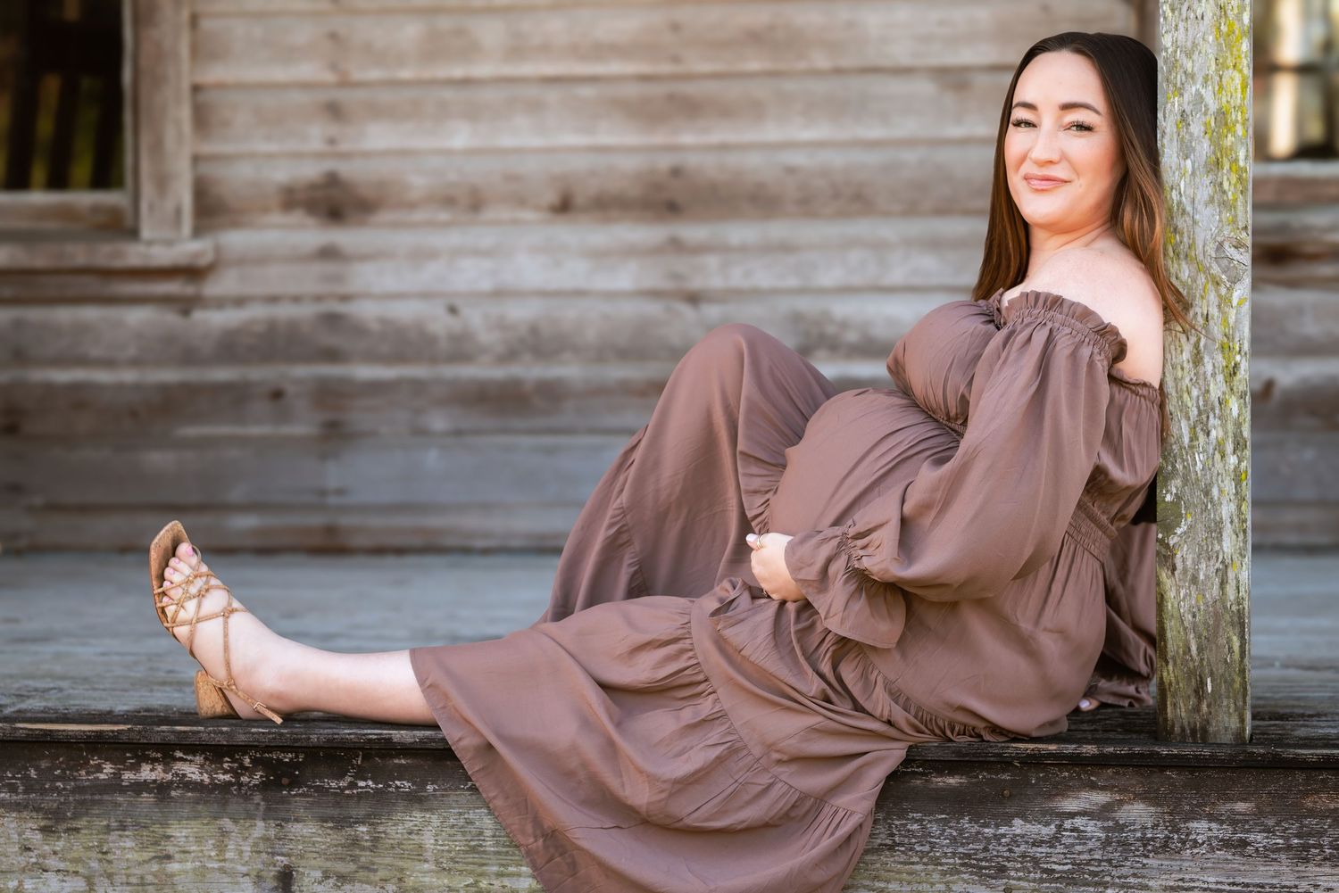 Young woman in brown off-shoulder maxi dress sitting on wooden steps leaning against a rustic wall.