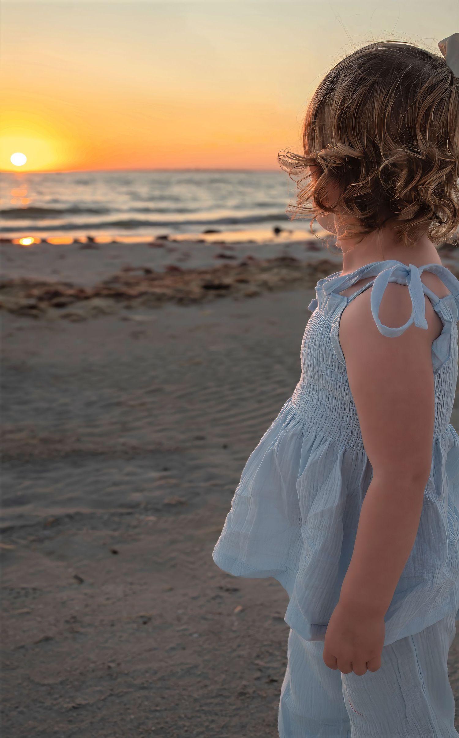 Young girl in white smocked dress watching a golden sunset over the ocean on a sandy beach.