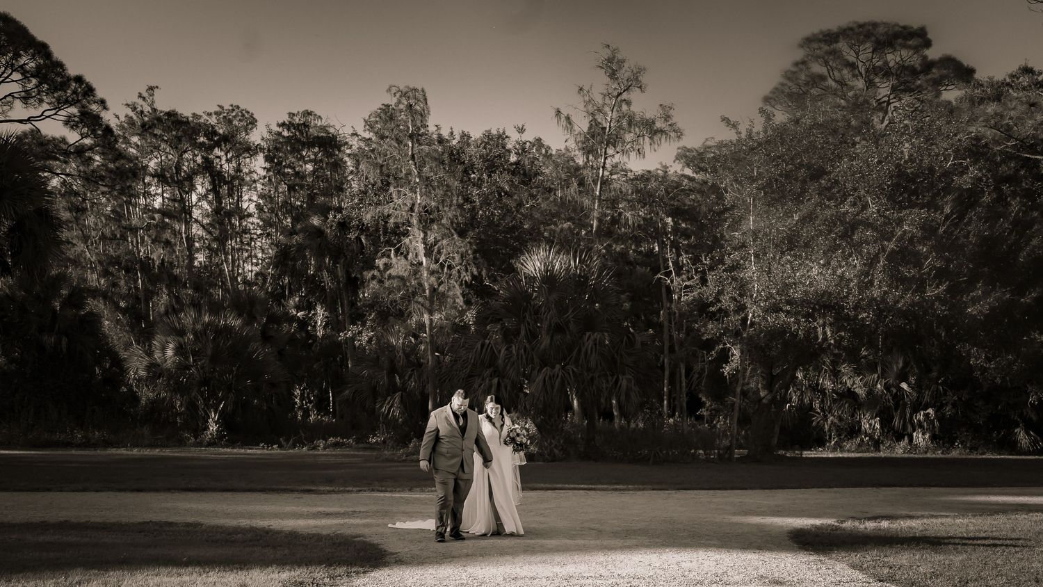 Black and white photo of bride and groom standing together in an open field surrounded by trees.