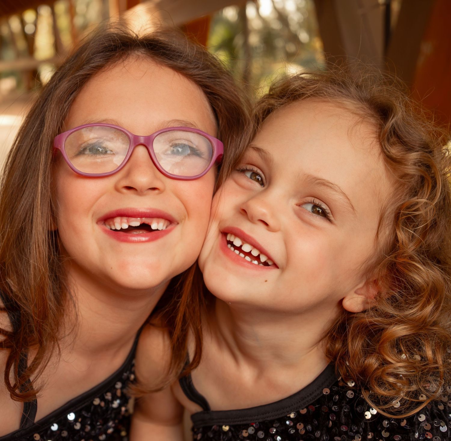 Close-up portrait of two smiling young sisters, one wearing pink glasses, cheek to cheek.
