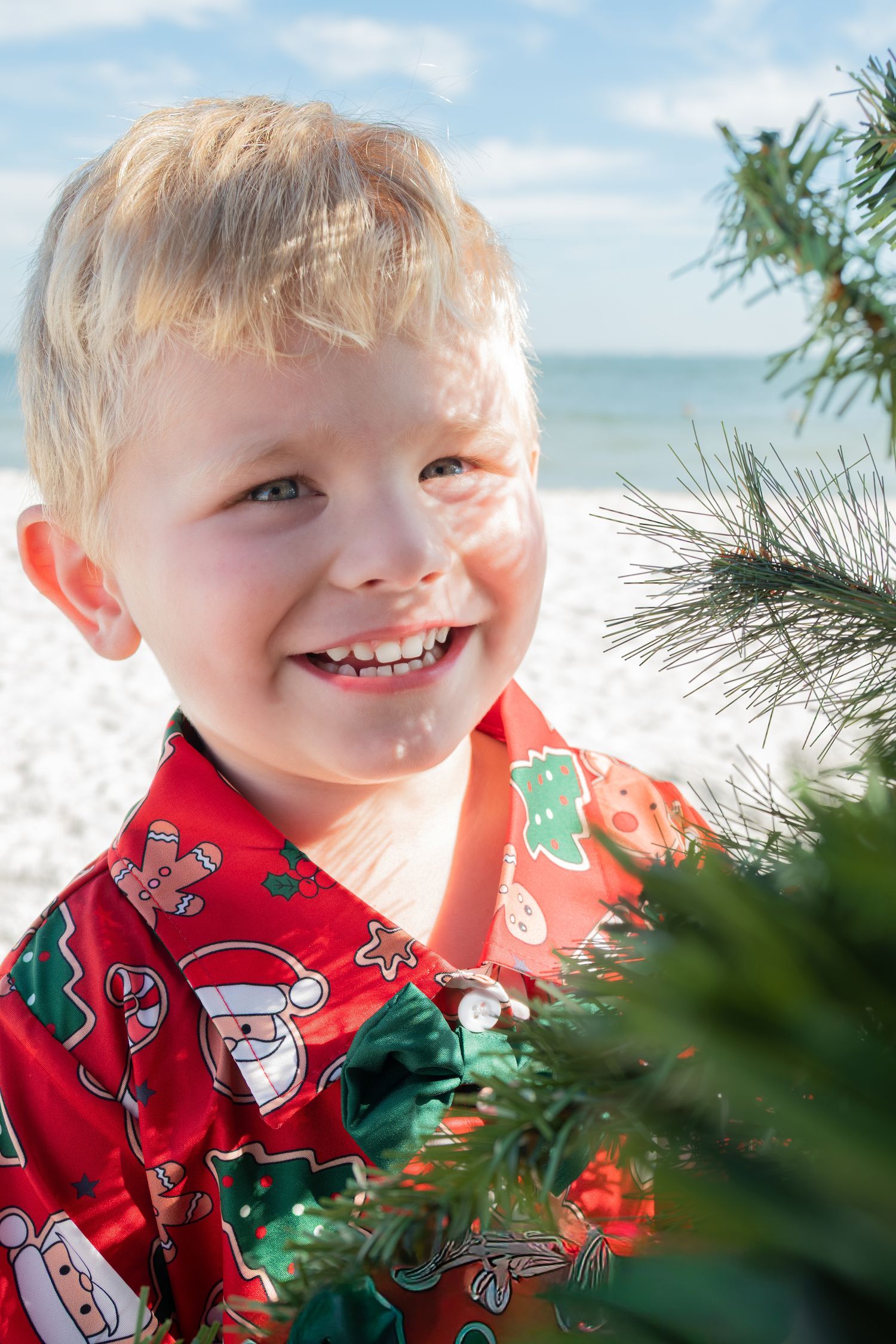 Smiling blonde boy in Christmas shirt near a decorated tree on the beach.