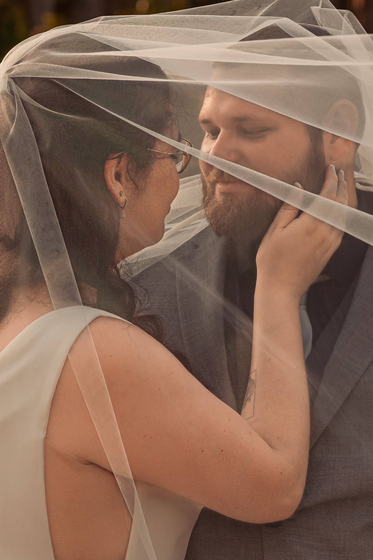 Bride and groom share an intimate moment together beneath a flowing bridal veil in warm light.