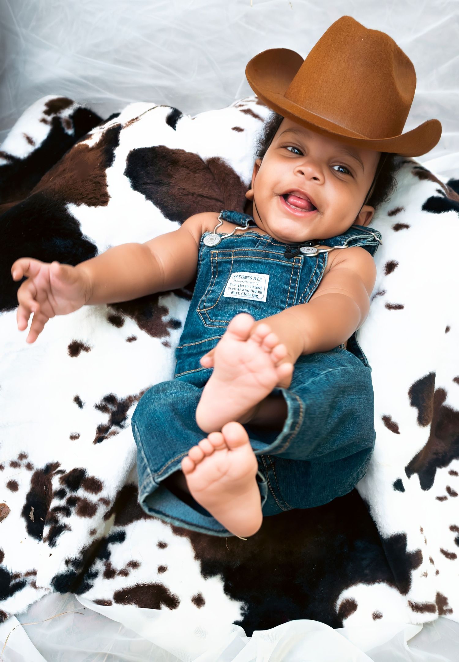 Adorable baby in denim overalls and brown cowboy hat smiling on cow print blanket.