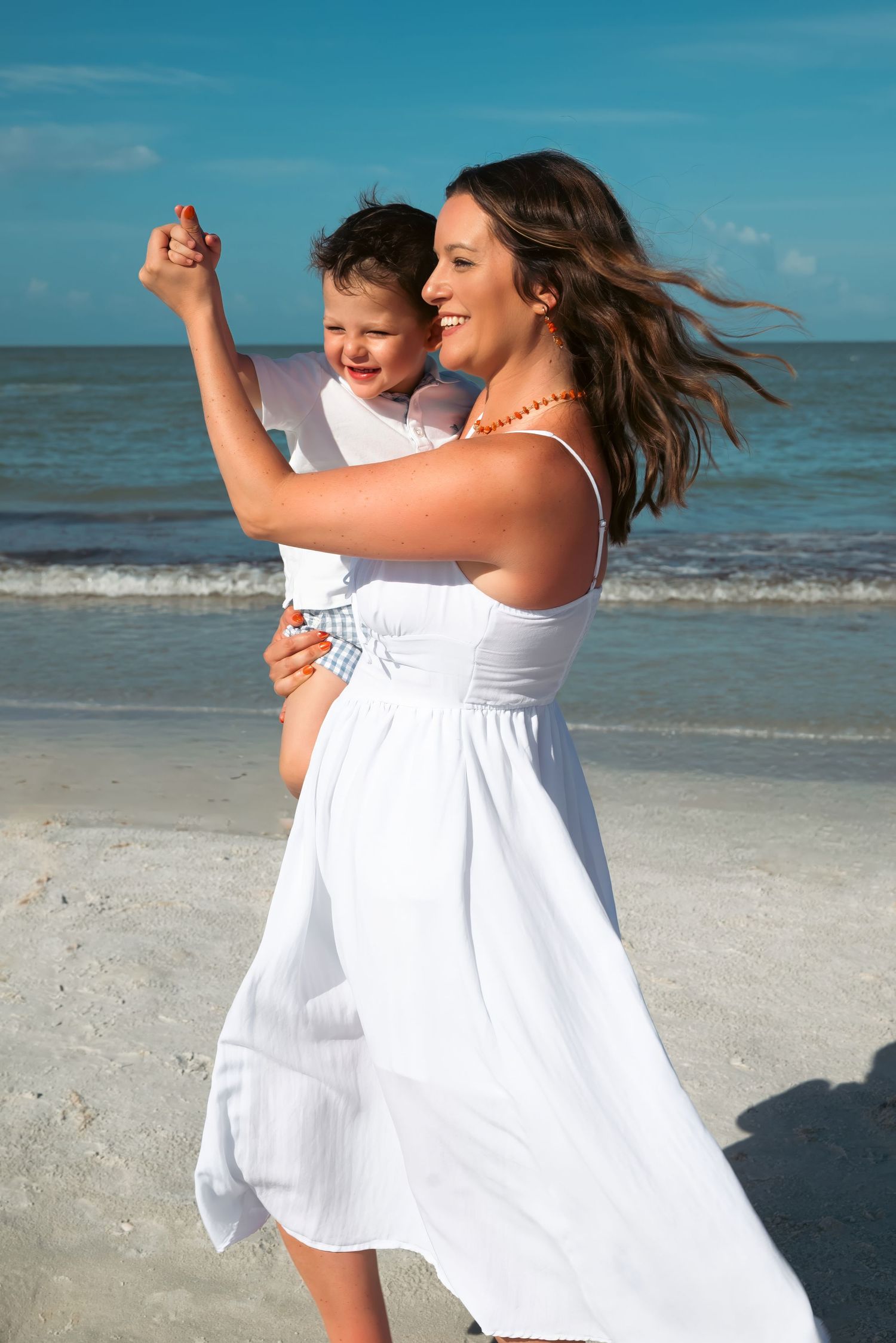 A smiling mother in a white dress holds her young child on a sunny beach with turquoise ocean waves.