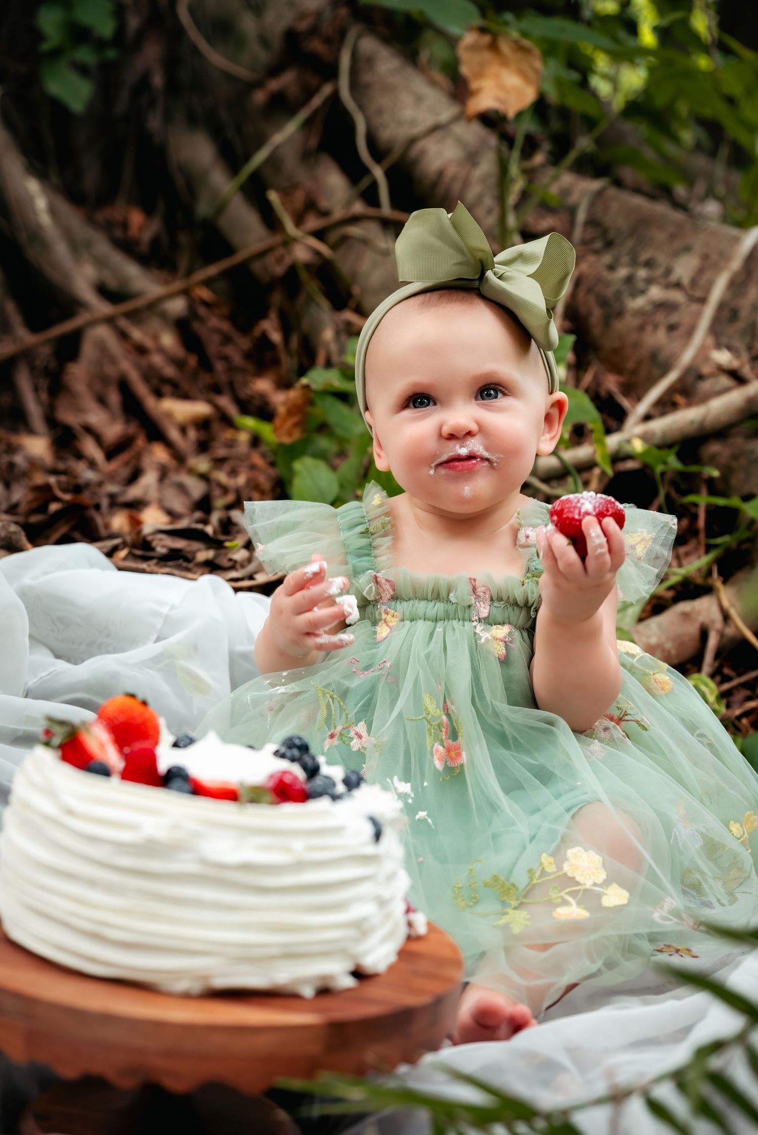 Baby girl in green tulle dress holds strawberry beside white cake with berries during outdoor cake smash photoshoot.