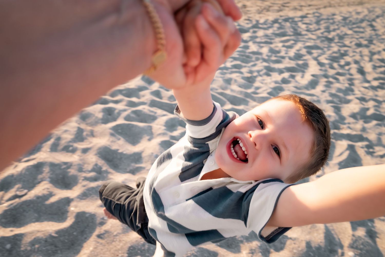 Happy boy being swung by an adult hand on a sandy beach, laughing joyfully in a striped shirt.