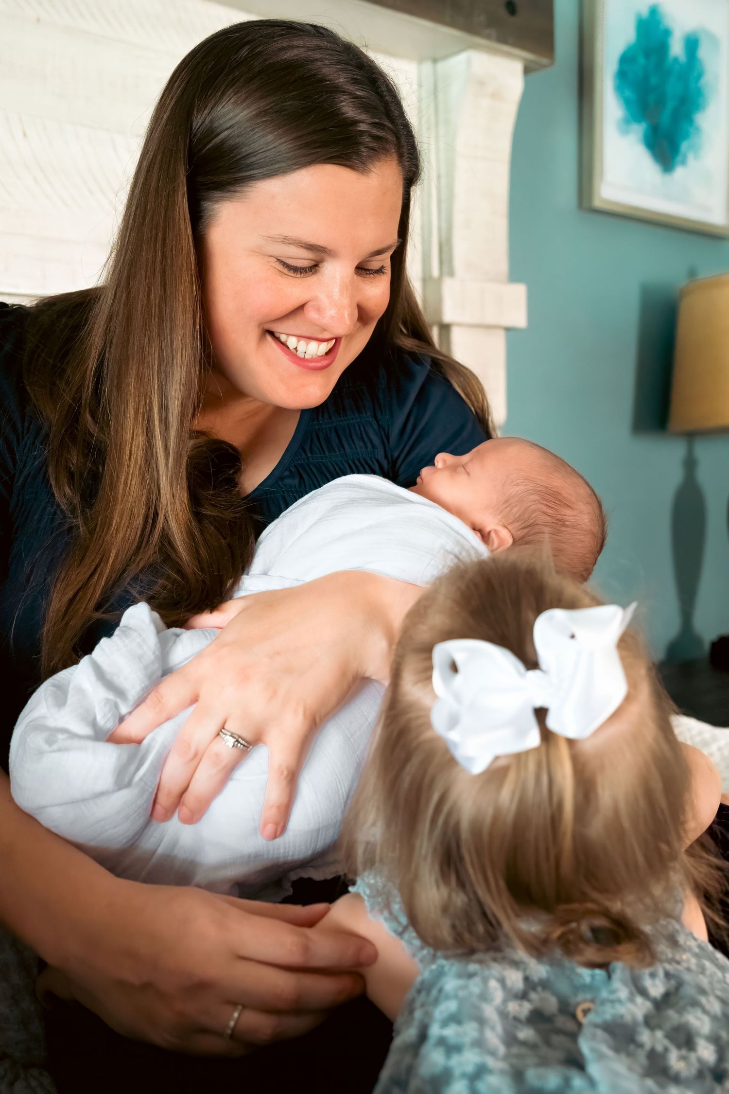 Smiling mother holding newborn baby while young toddler girl with white bow looks on indoors.