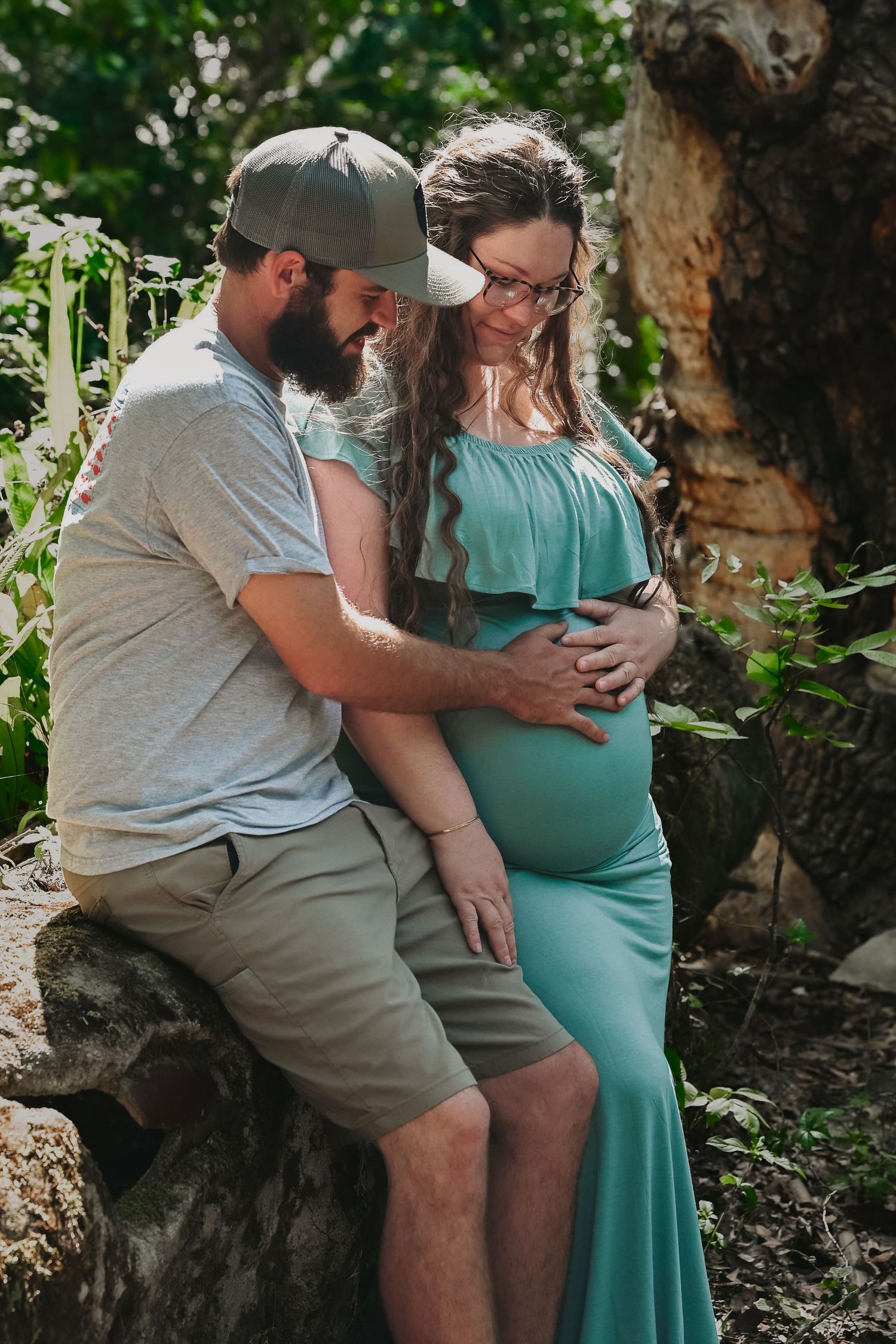 Bearded man tenderly holding pregnant woman's belly while sitting on rocks in lush greenery.
