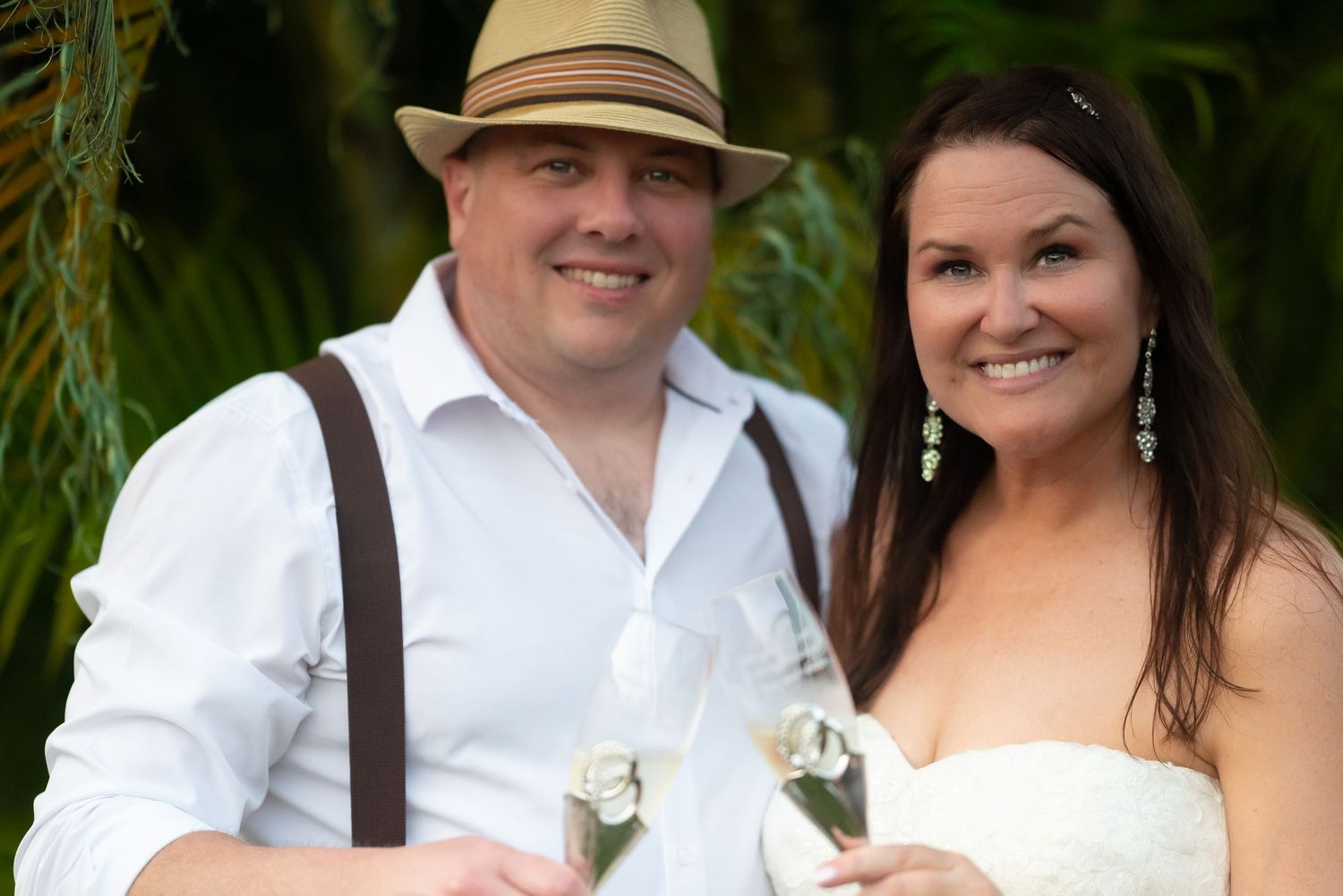 Smiling bride and groom pose together outdoors, groom wearing suspenders and straw hat, bride in white strapless dress.