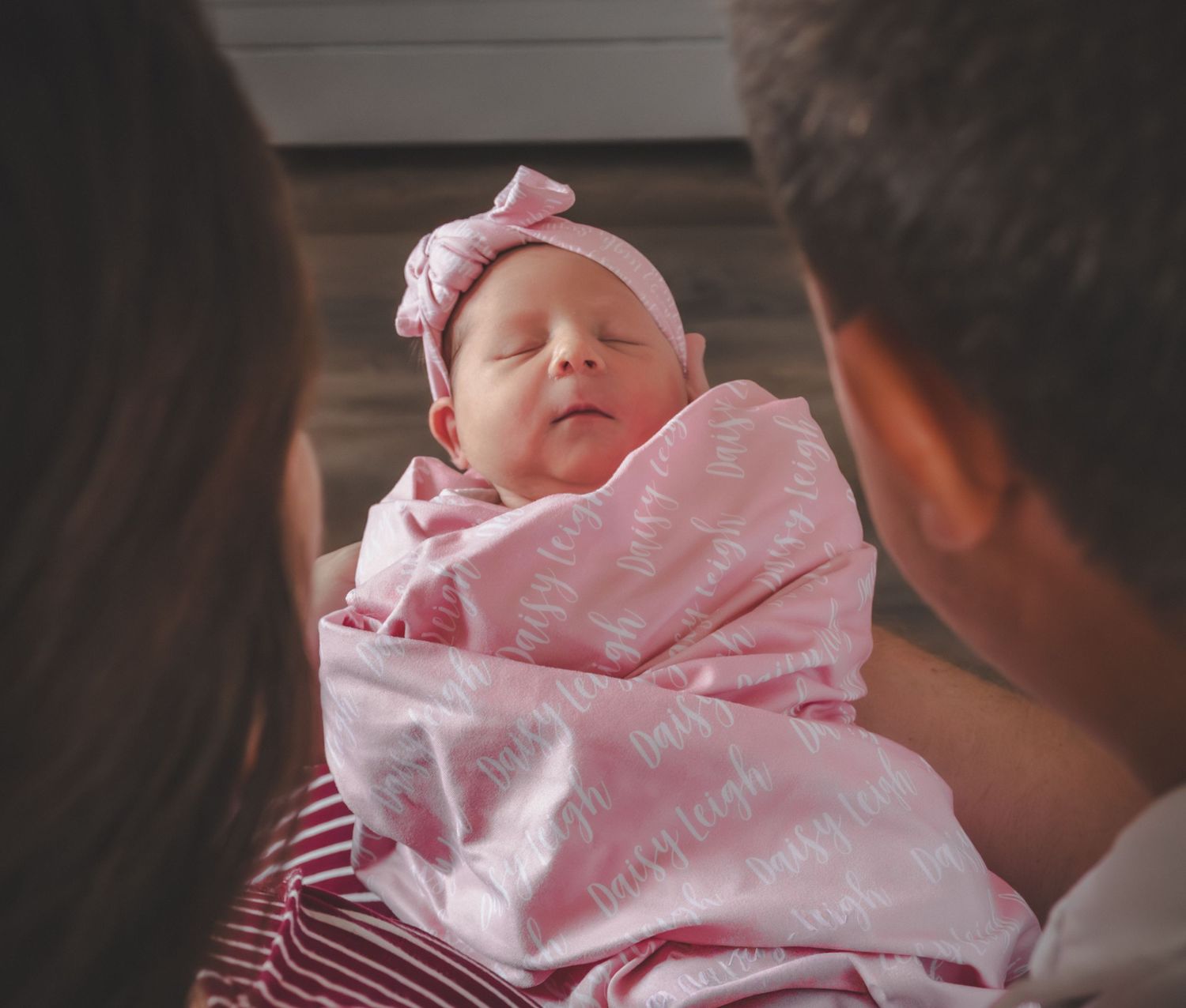 Parents lovingly hold sleeping newborn baby girl in pink bow headband and swaddle.