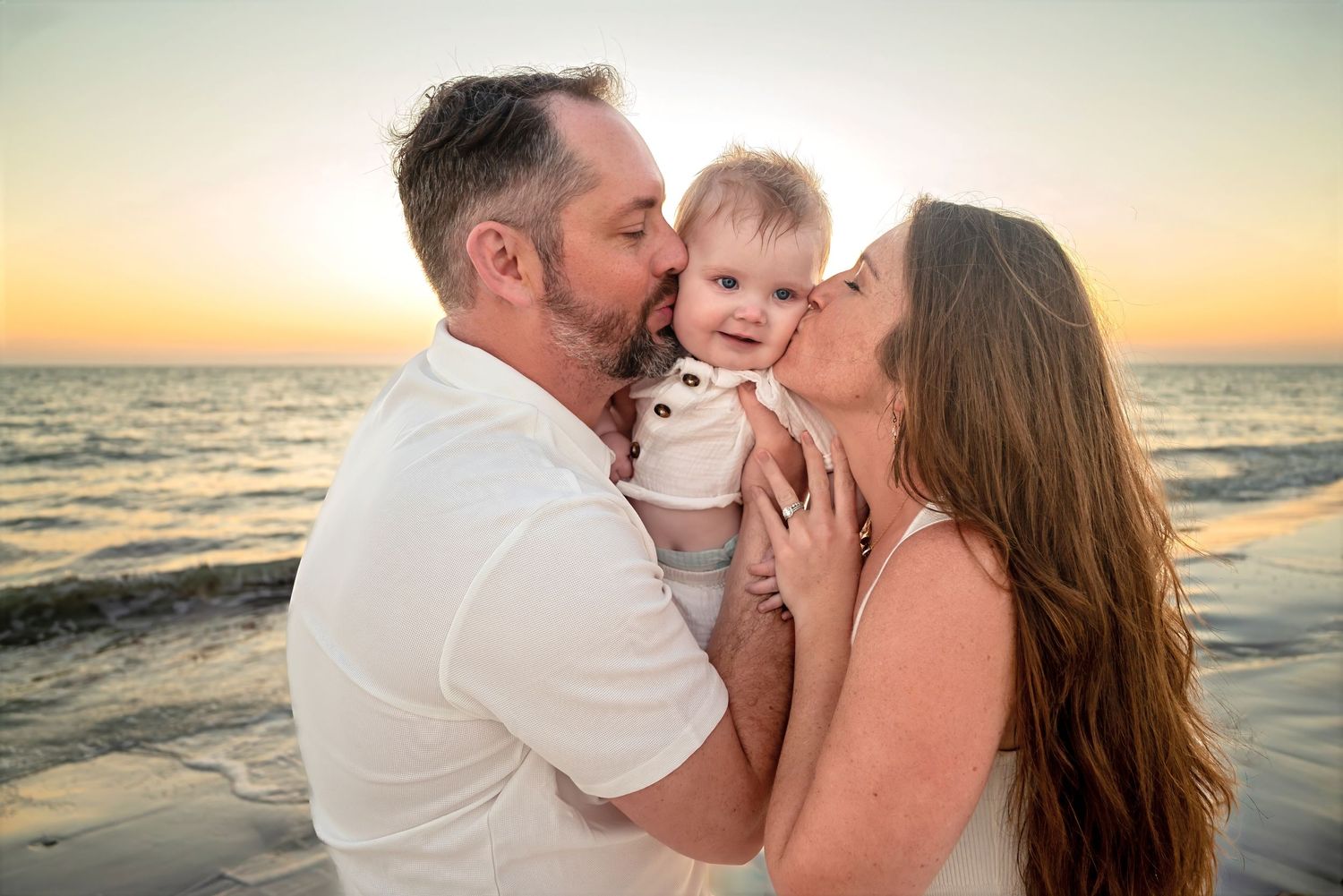 Happy couple kissing smiling baby on cheeks simultaneously at sunset on the beach.