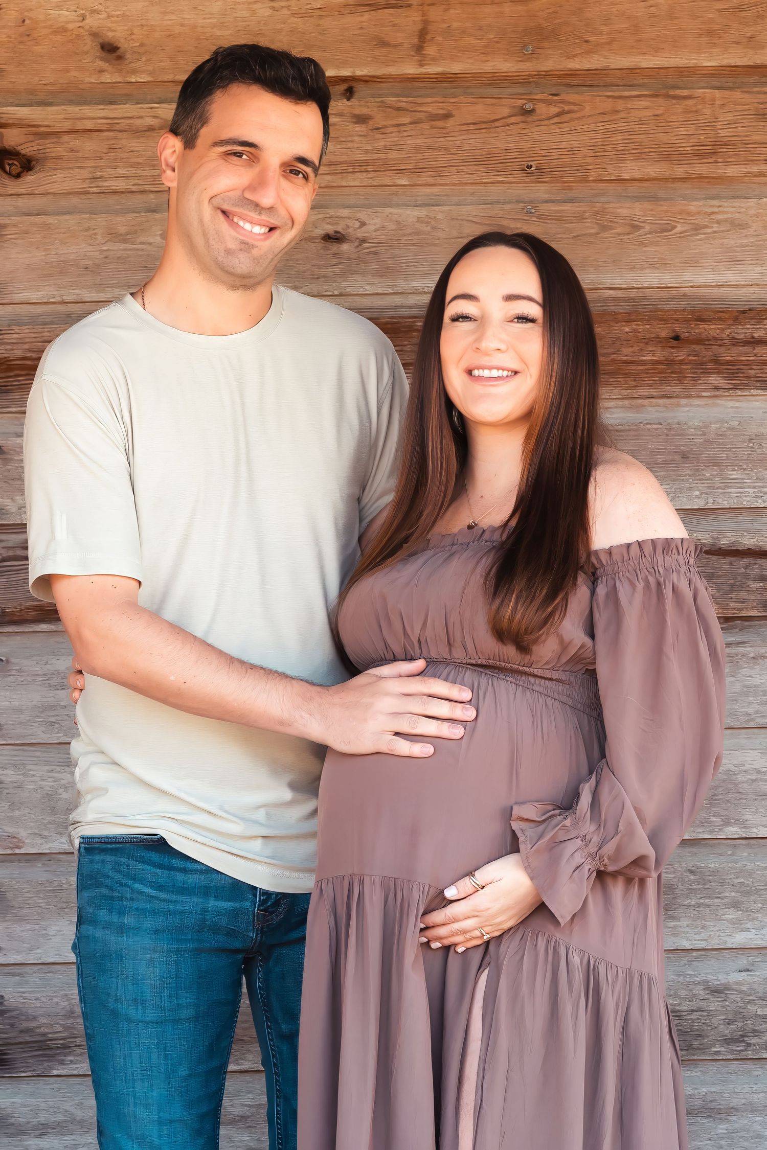 Smiling expecting couple posing together in front of a rustic wooden log wall for maternity portrait.