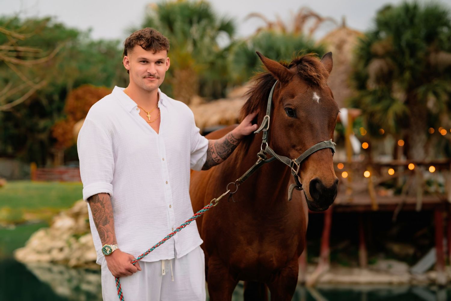 Handsome tattooed groom in white linen outfit stands beside a chestnut horse at an outdoor wedding venue.