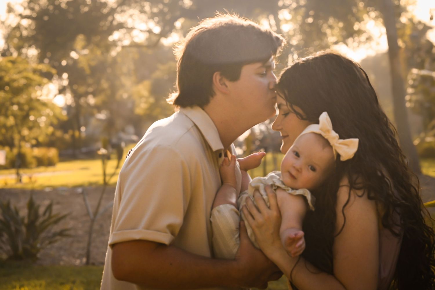 Young couple sharing a kiss over their baby in warm golden sunlight outdoors.