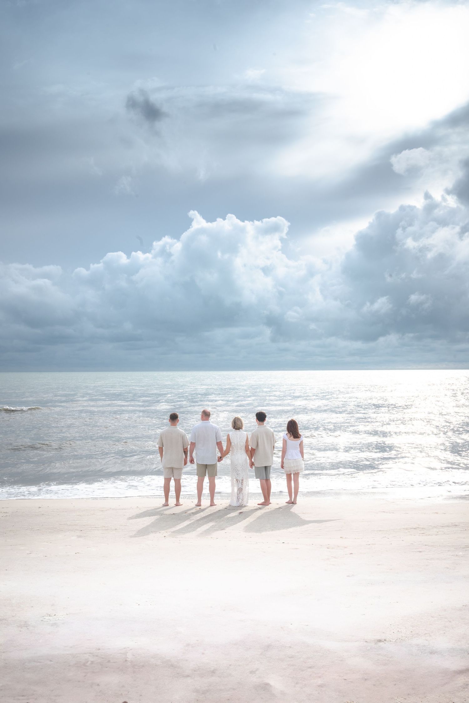 Family of five standing together on sandy beach gazing at dramatic cloudy ocean horizon.