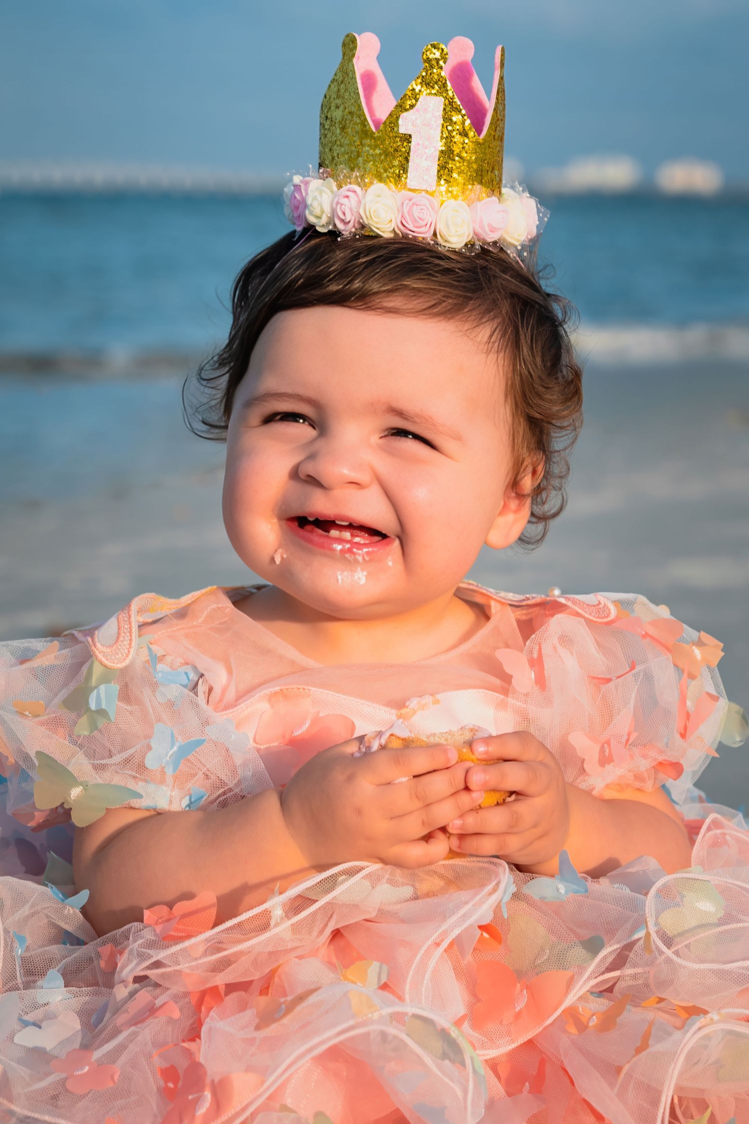 Happy baby girl celebrating first birthday at beach in pink dress and gold crown.