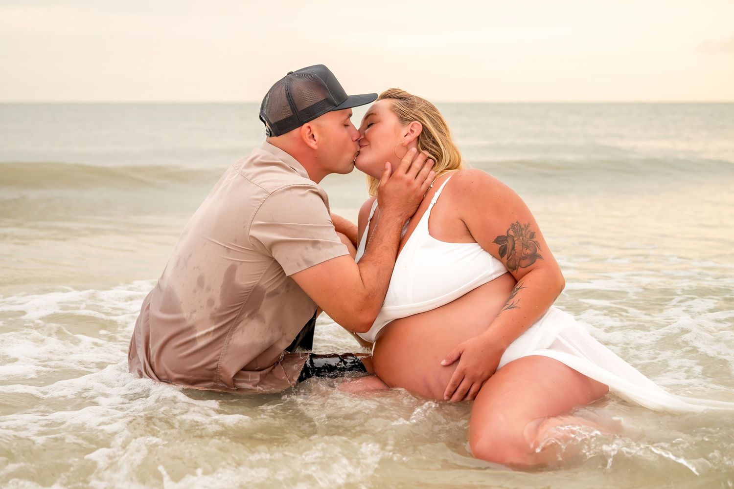 Pregnant couple sharing a romantic kiss while sitting in ocean waves during a beach maternity photo shoot.