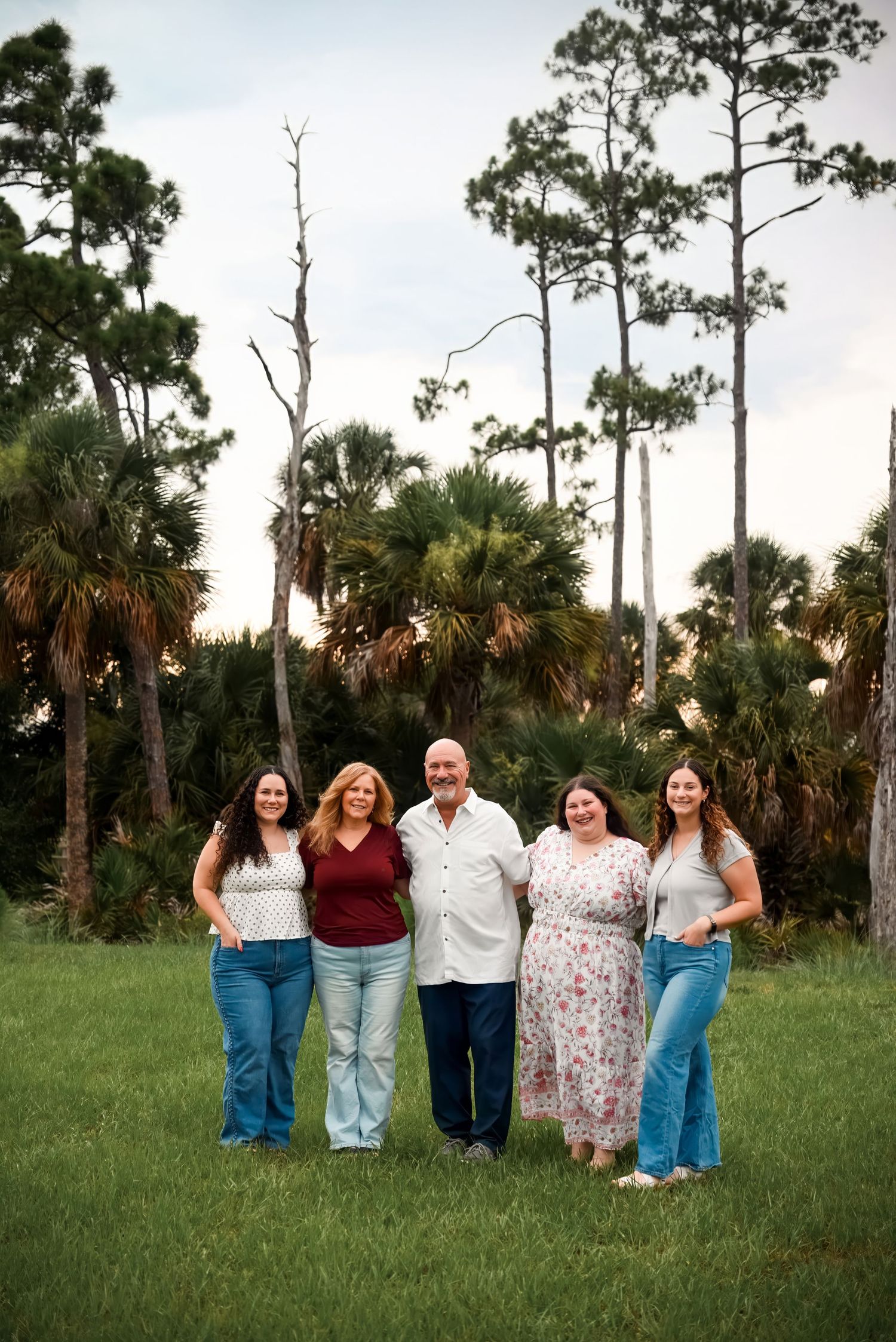 Family of five smiling together outdoors on green grass with palm trees in background during golden hour.