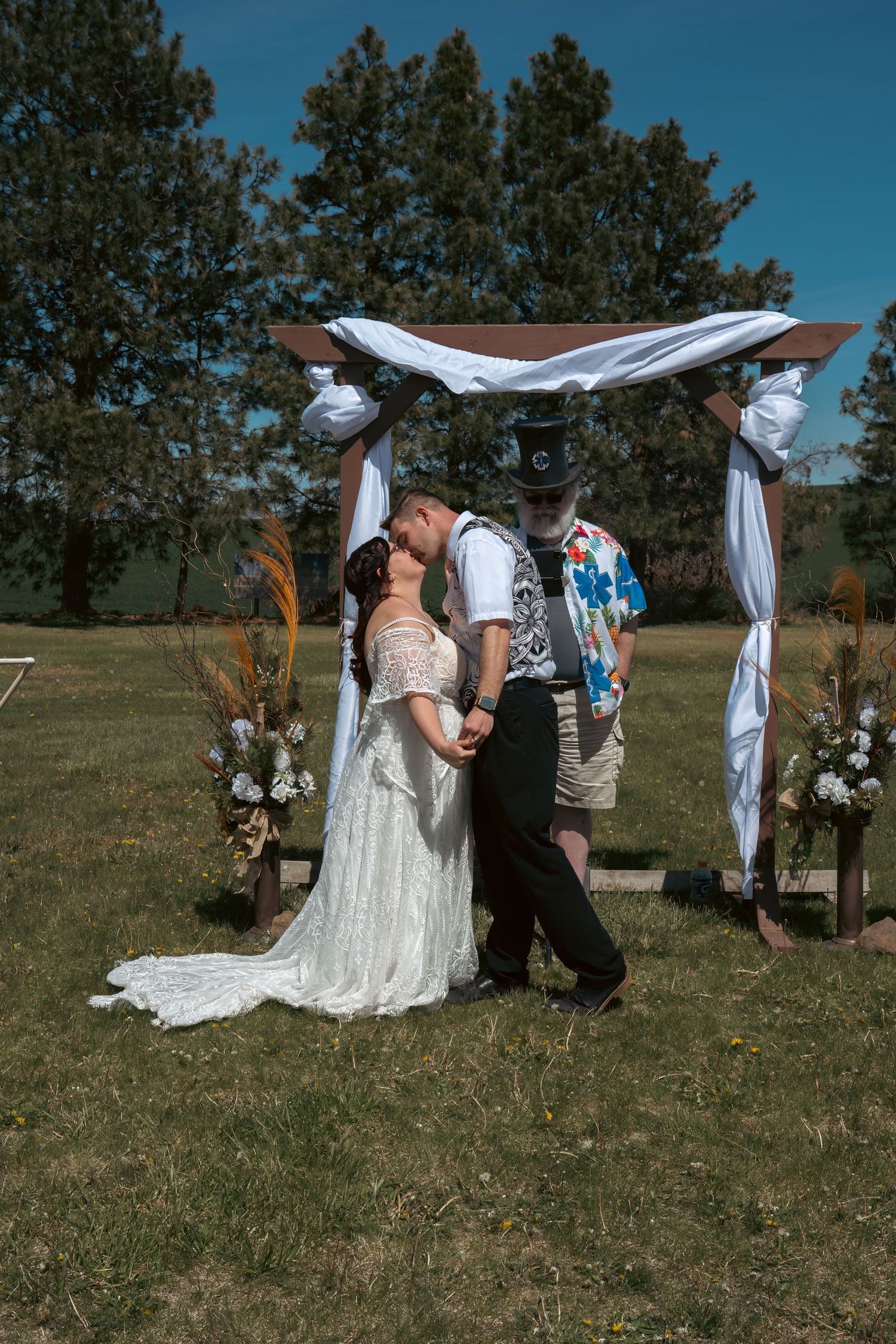 Bride and groom share first kiss under white draped wedding arch outdoors with tall pine trees and blue sky.