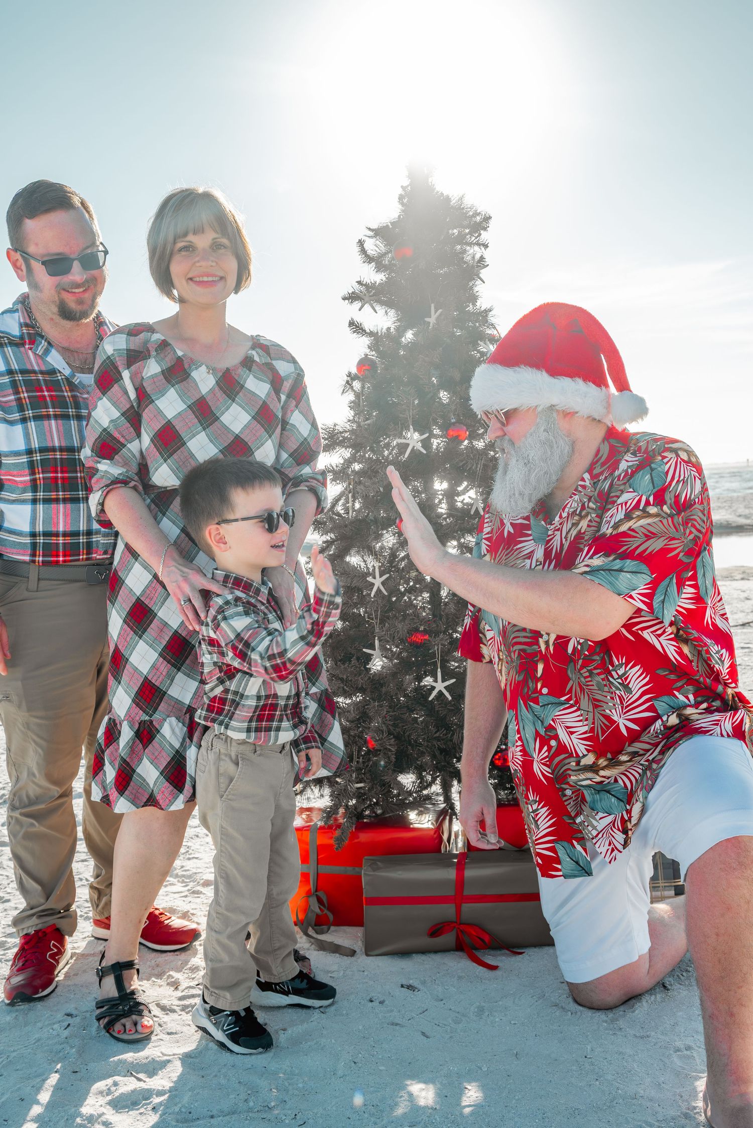 Beach Santa Claus gives high five to young boy in sunglasses while family watches during Christmas beach photo shoot.