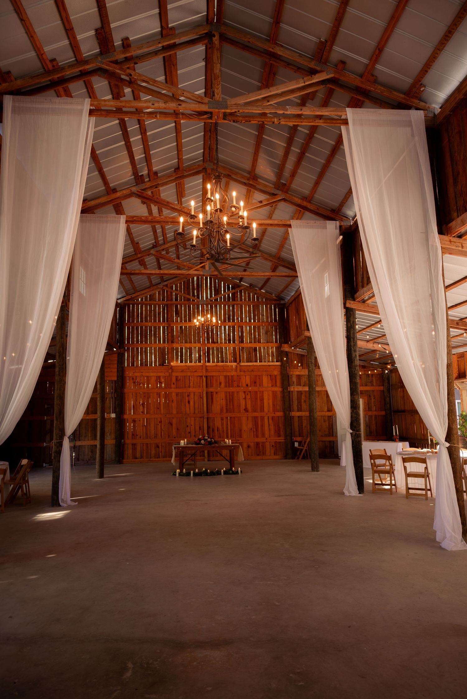 Rustic barn wedding venue interior featuring white draping curtains, wooden ceiling beams, and warm chandelier lighting.