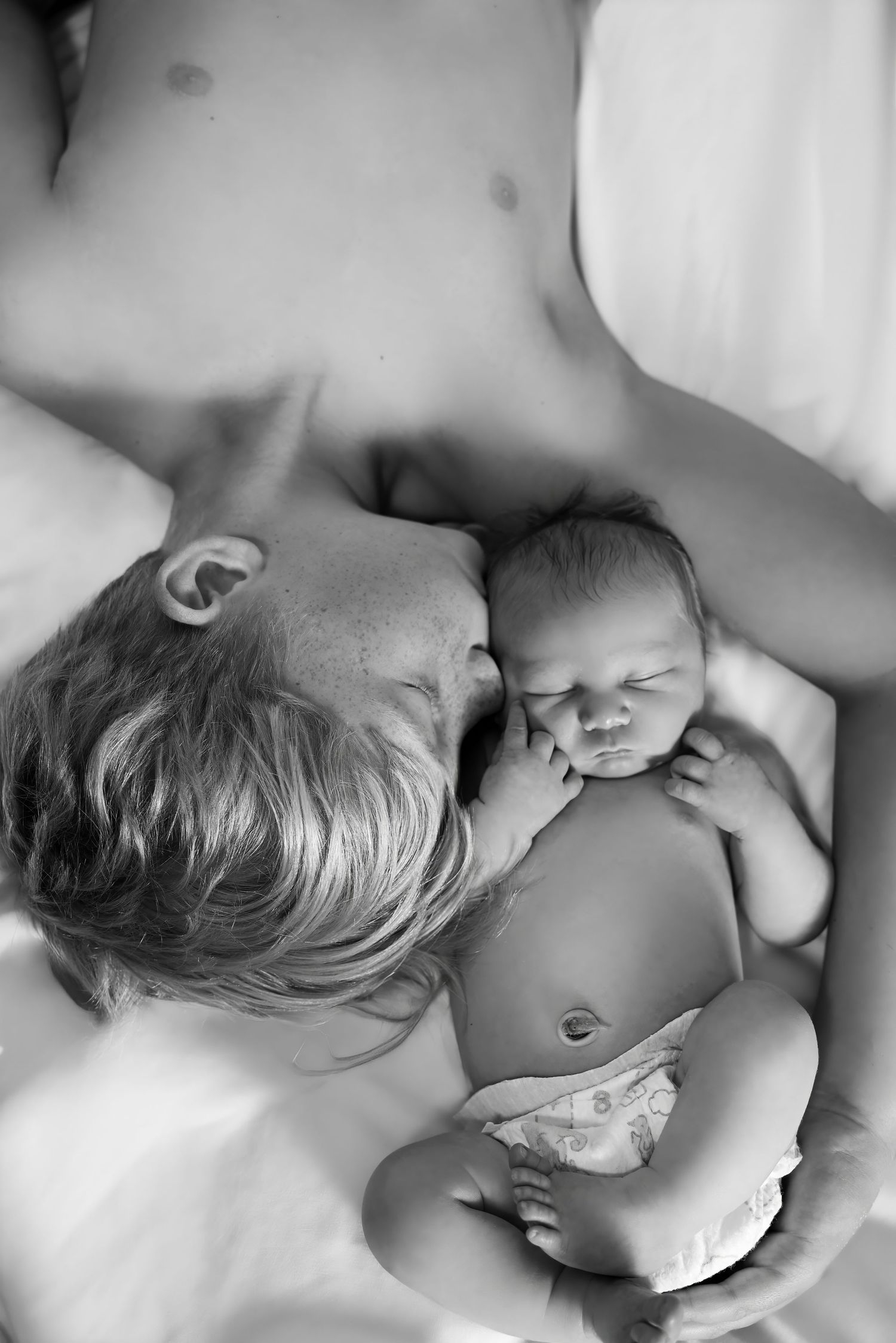 Black and white photo of a young child tenderly resting beside a sleeping newborn baby on white bedding.