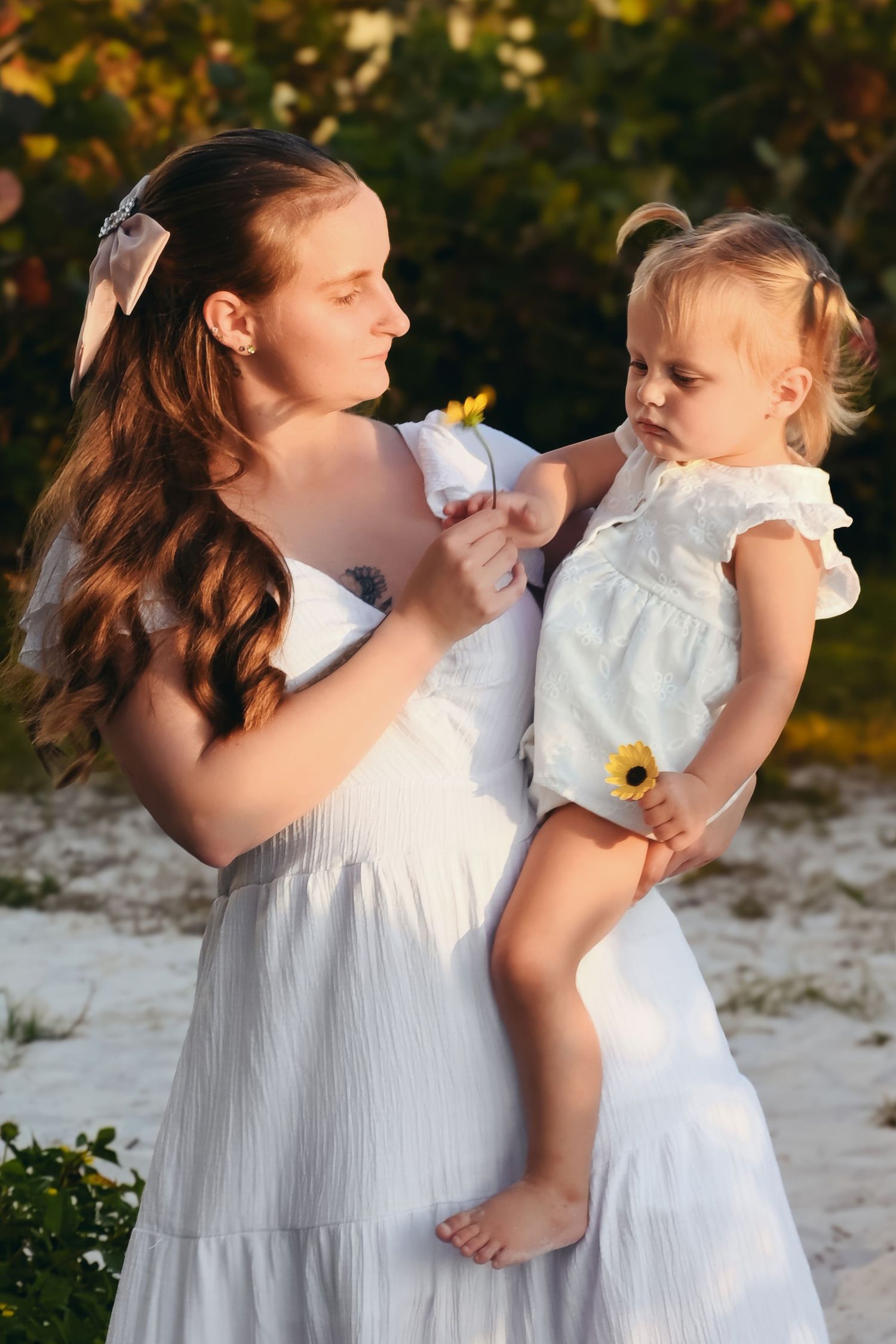 A young woman in a white dress holds a toddler girl in white, both outdoors at golden hour with yellow flowers.