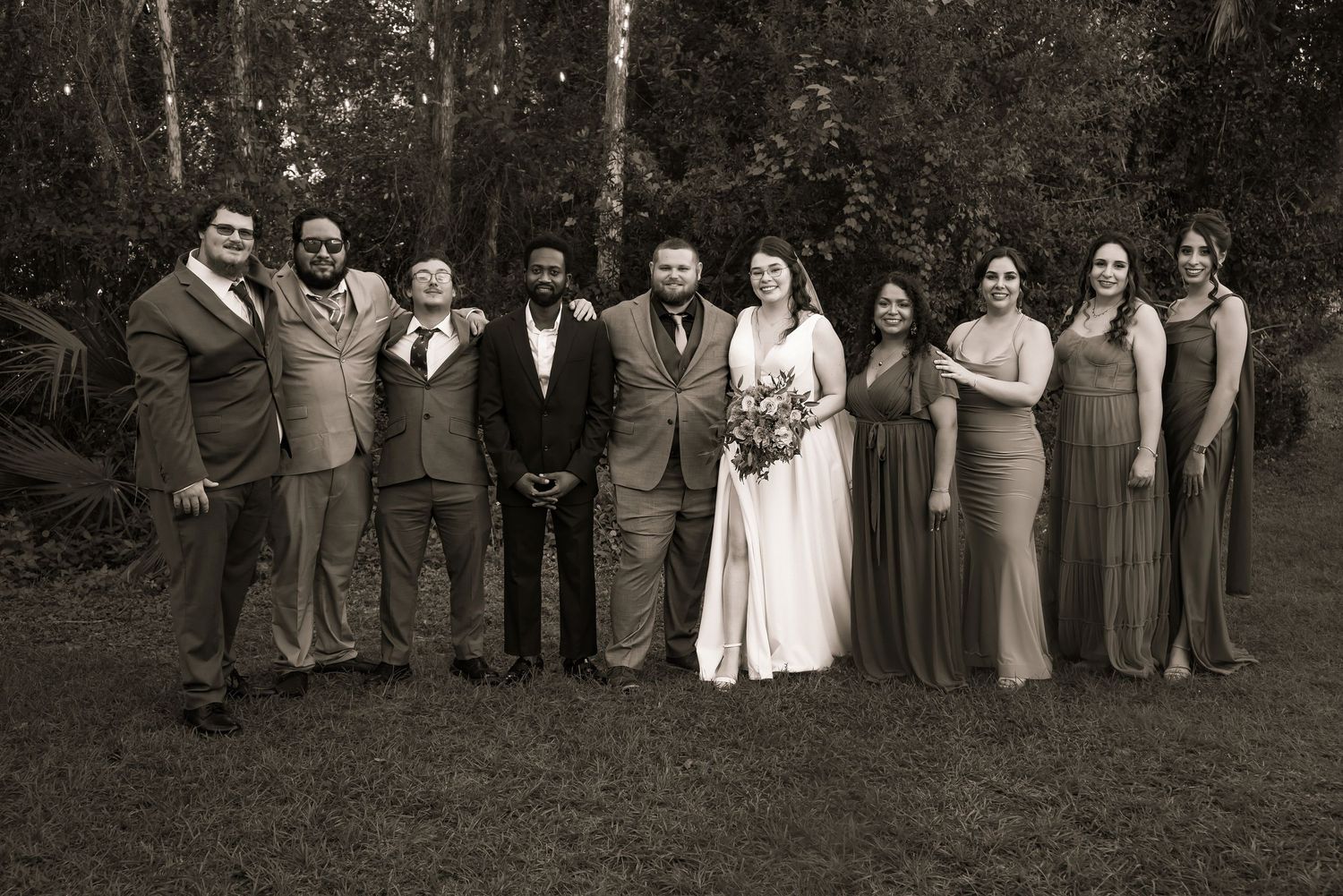 Black and white wedding party portrait with bride, groom, groomsmen, and bridesmaids in nature.