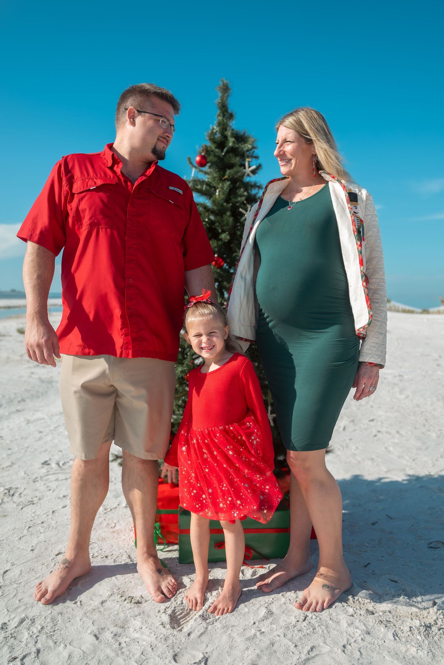 Pregnant family of three in Christmas outfits posing by a beach Christmas tree.