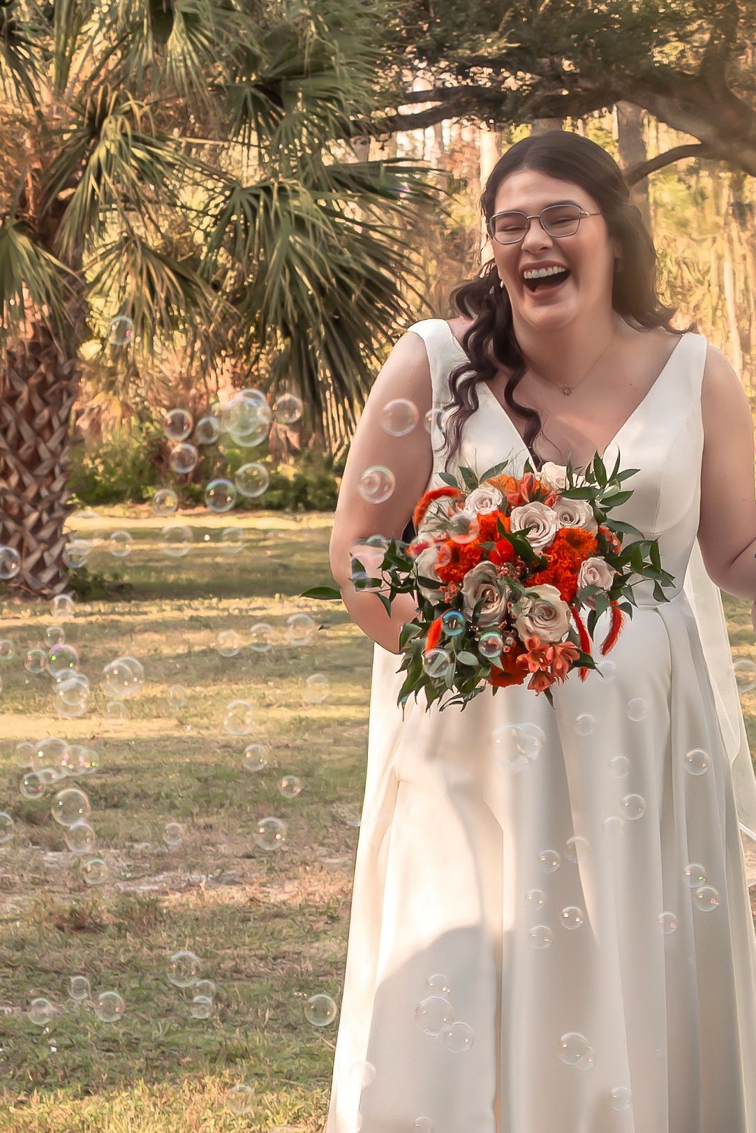 Joyful bride in white gown holding orange floral bouquet laughing among floating bubbles outdoors.