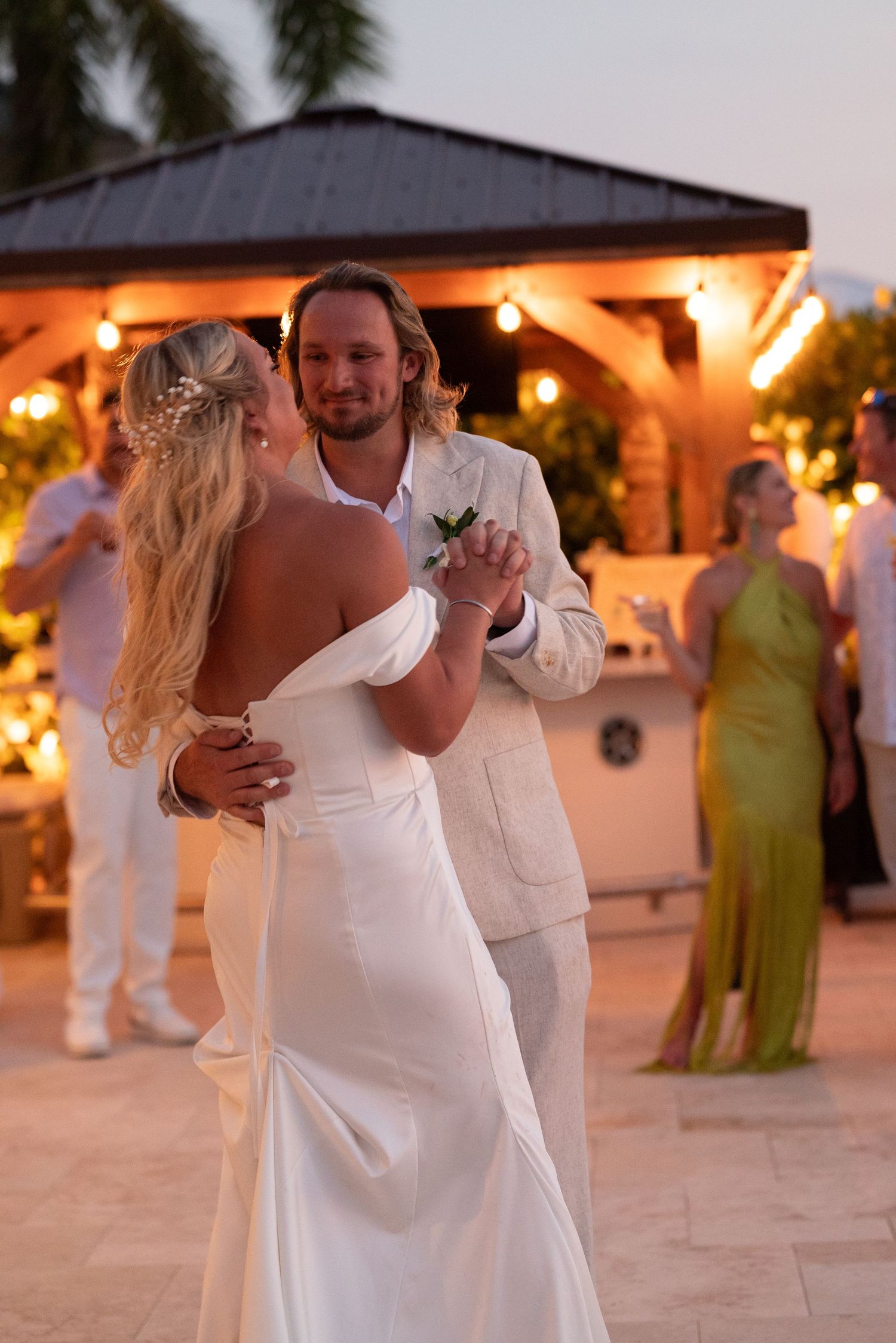 Bride and groom share first dance outdoors at sunset wedding reception with string lights and guests in background.